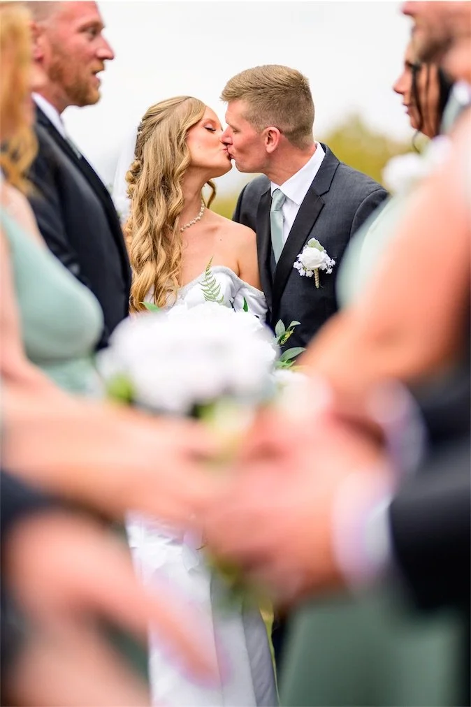 Bride and groom share a kiss surrounded by guests with blurred hands in the foreground
