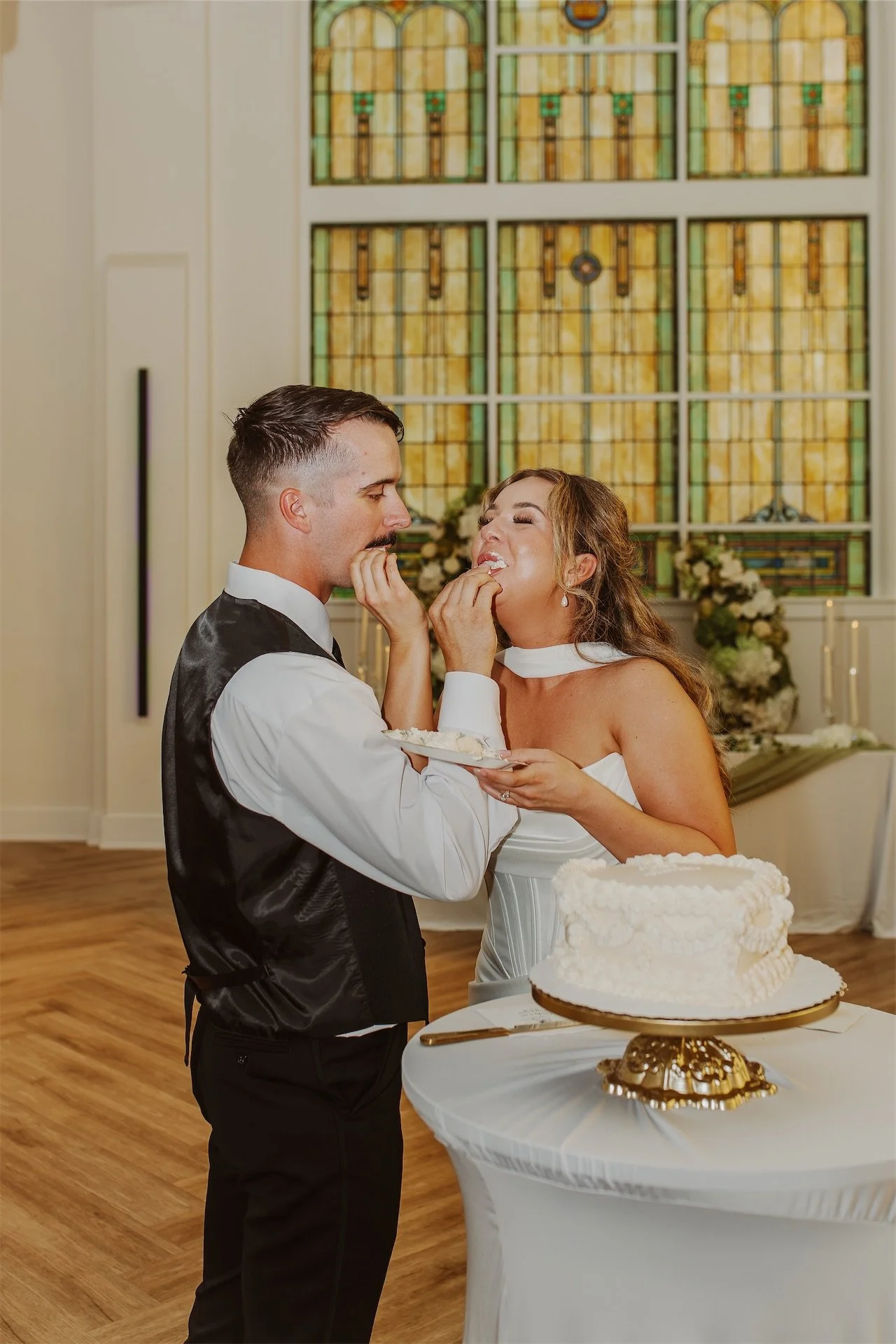 Bride and groom feeding each other wedding cake beside stained glass window