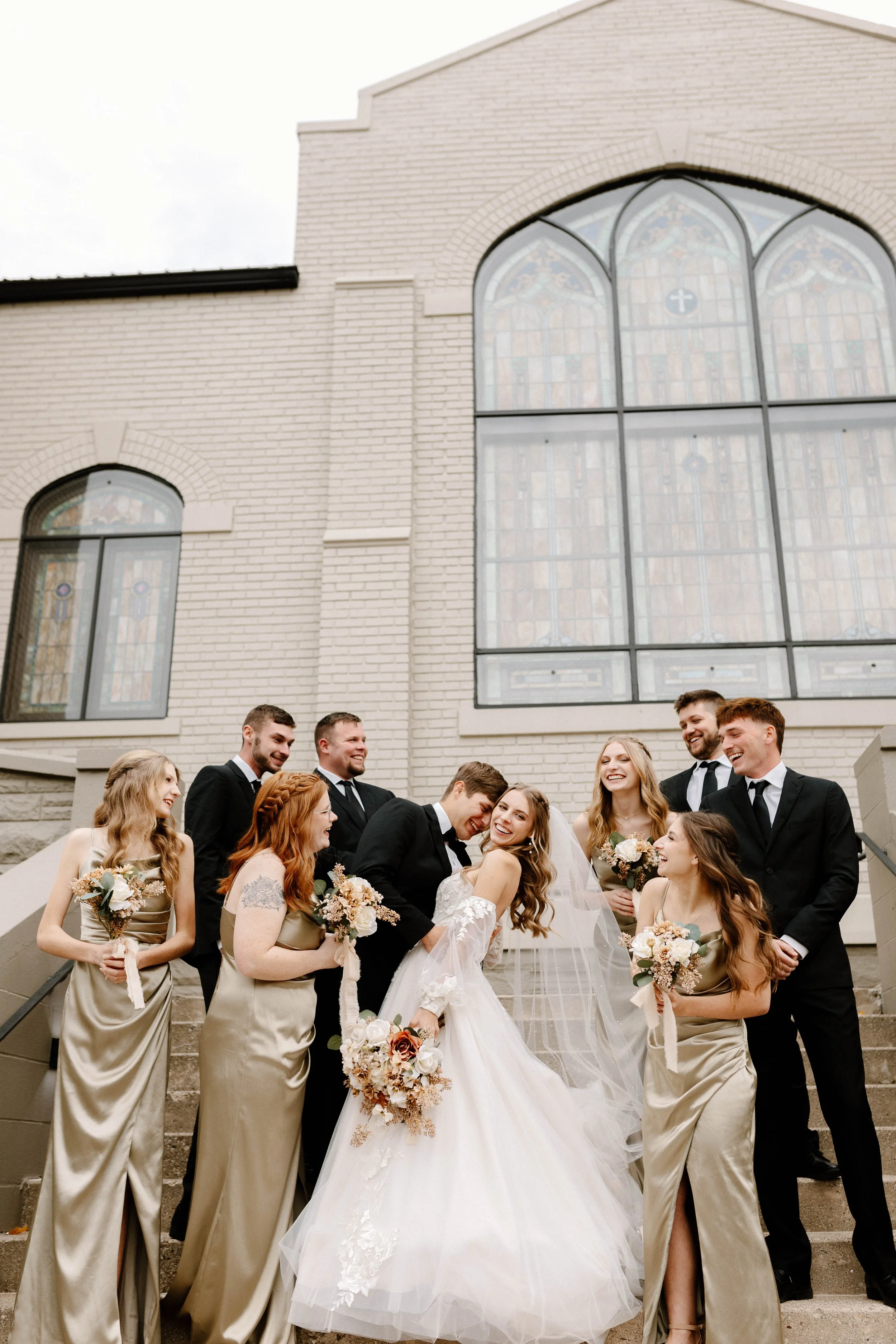 A wedding party standing on church steps outside, with a bride and groom in the center, all smiling and holding bouquets, dressed in formal attire, in front of stained glass windows.