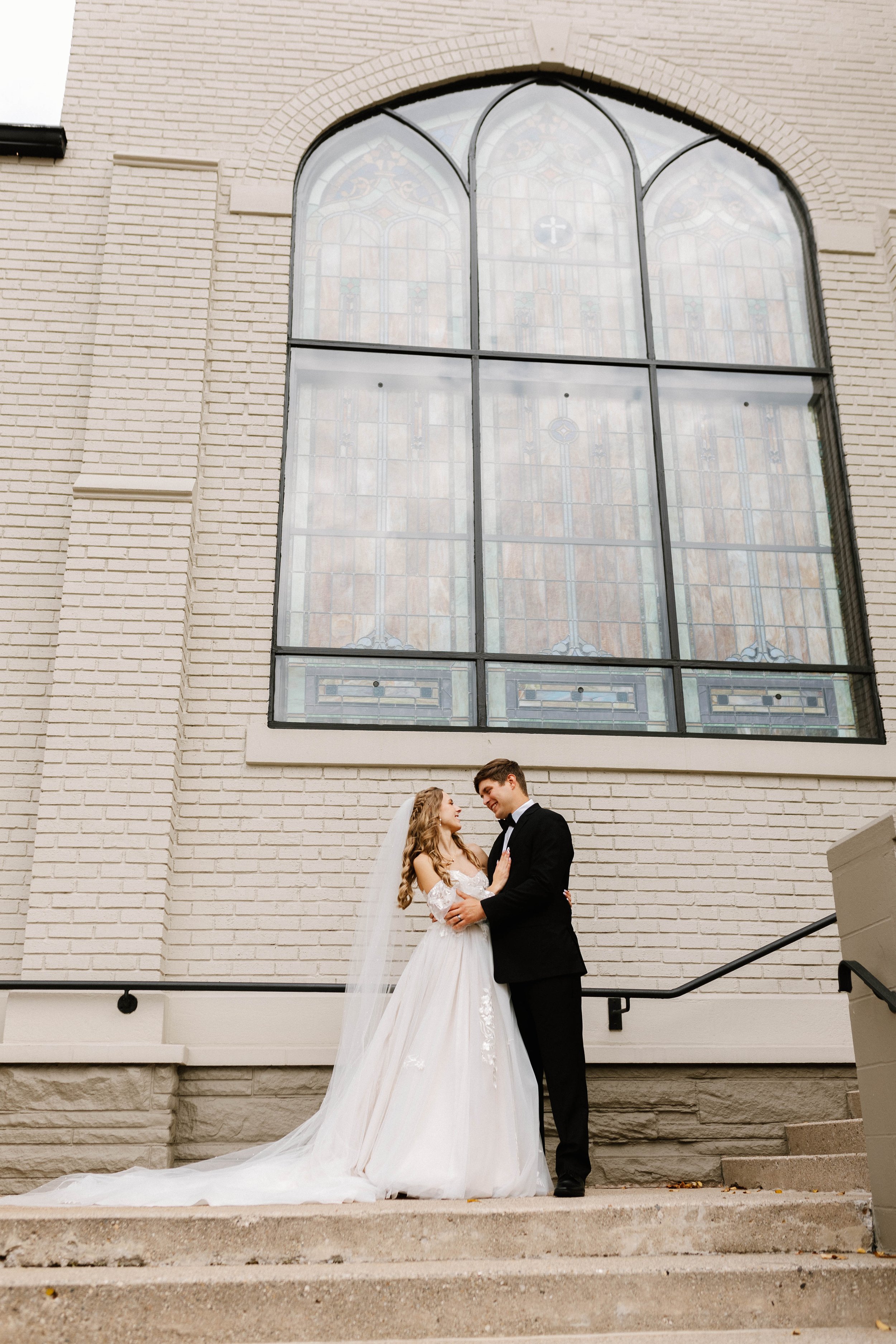 A bride in a white wedding gown and veil and a groom in a black tuxedo stand close together on steps outside a building with a large stained-glass window behind them.