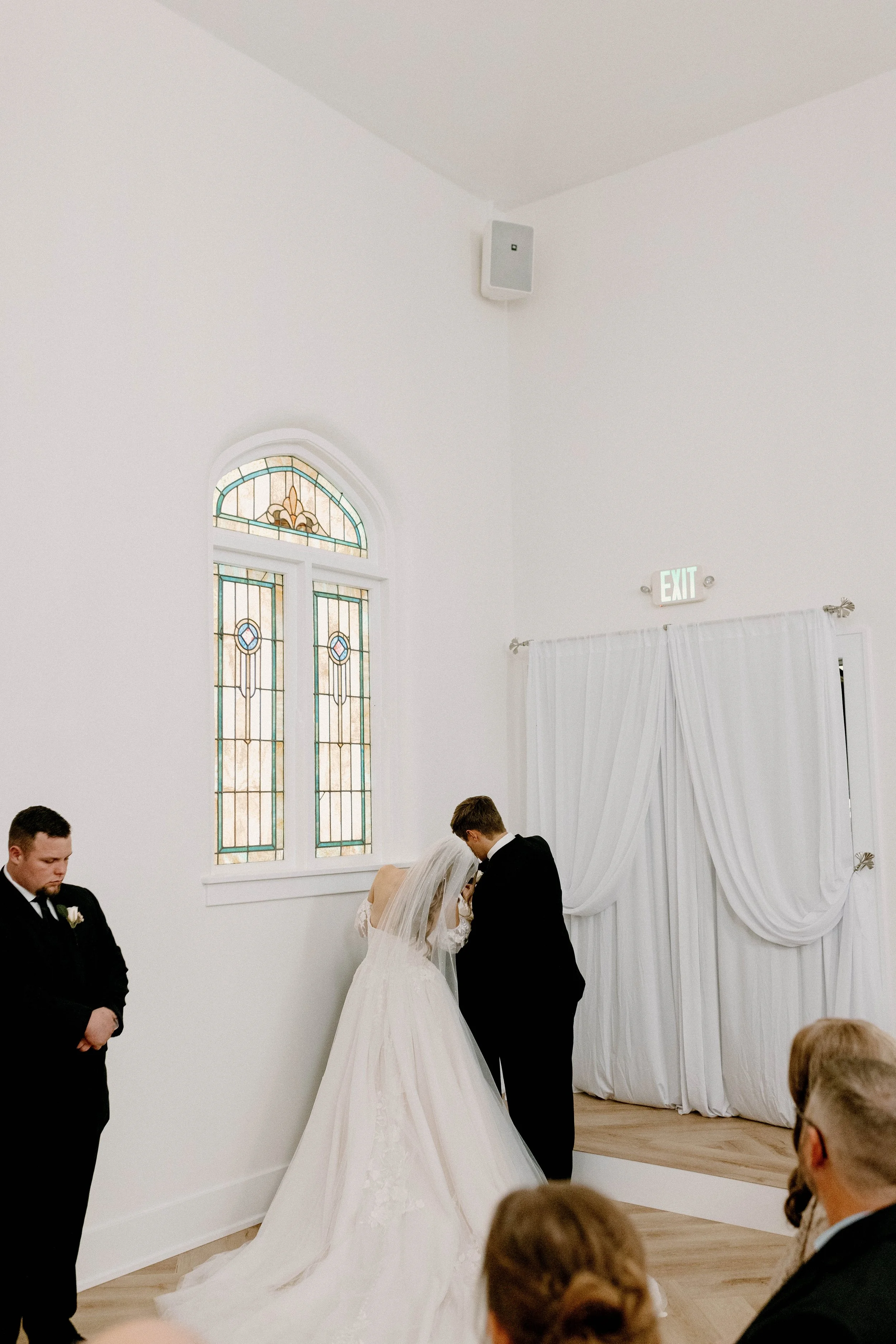 A bride and groom share a kiss during their wedding ceremony inside a white chapel with stained glass windows, while a man in a black suit stands nearby, and guests are seated watching.