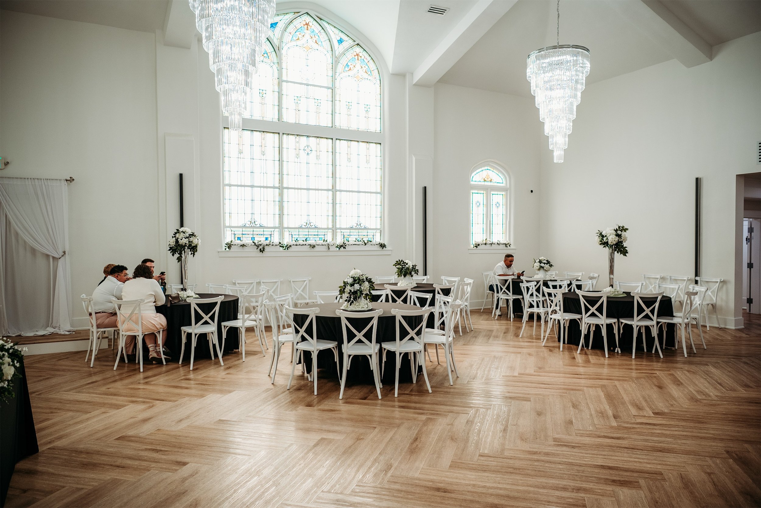 Reception hall with round black tables, white cross back chairs and crystal chandeliers beneath an arched stained glass window