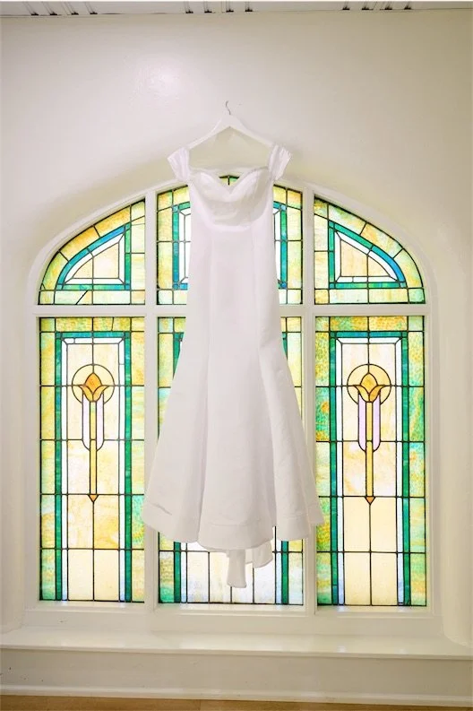 White wedding gown hanging from a hook in front of an arched stained glass window