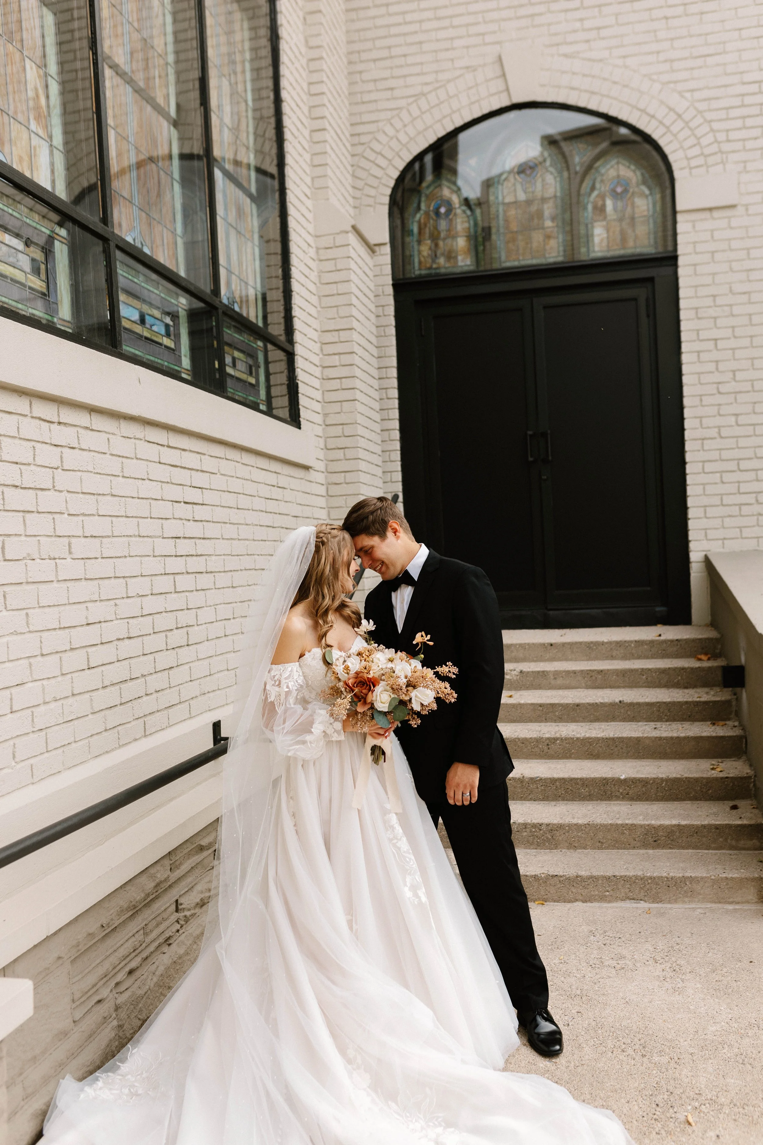 A bride and groom share a tender moment on their wedding day outside near stairs and a black door, with stained glass windows above.