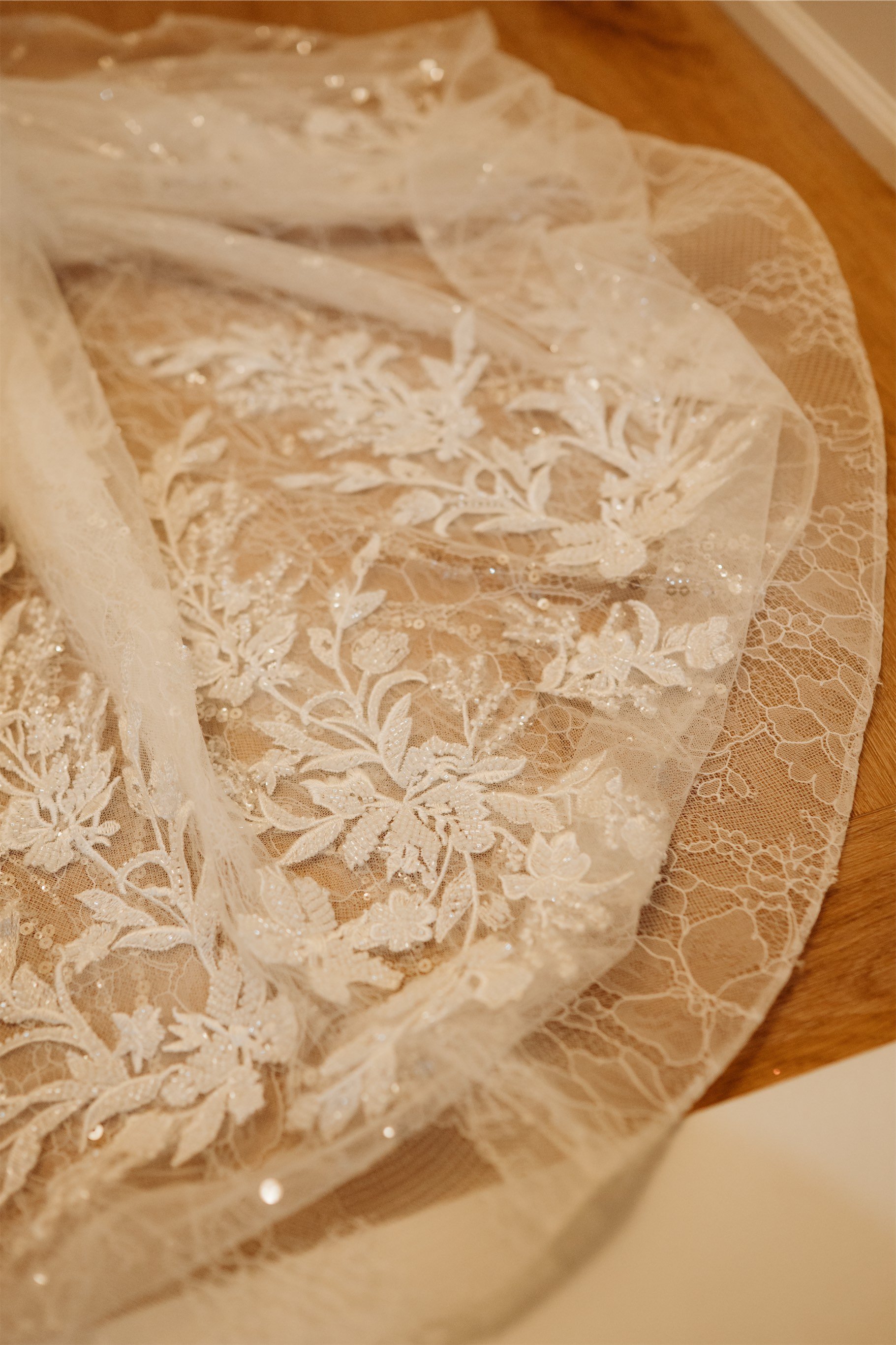Close up of floral lace embroidery and sequins on the bridal veil and gown train spread across a wood floor