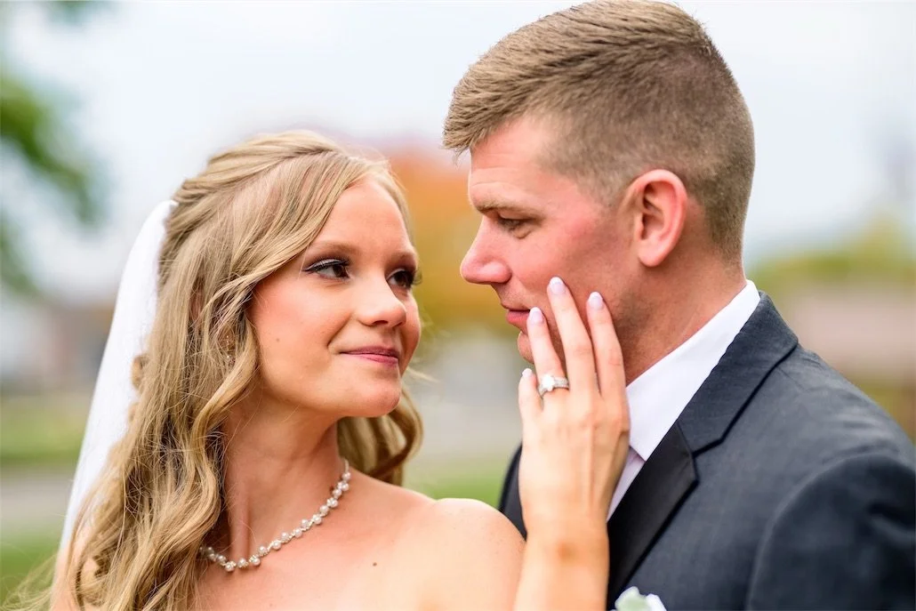 Bride gazes at her groom with her hand on his cheek, ring visible, outdoors in autumn