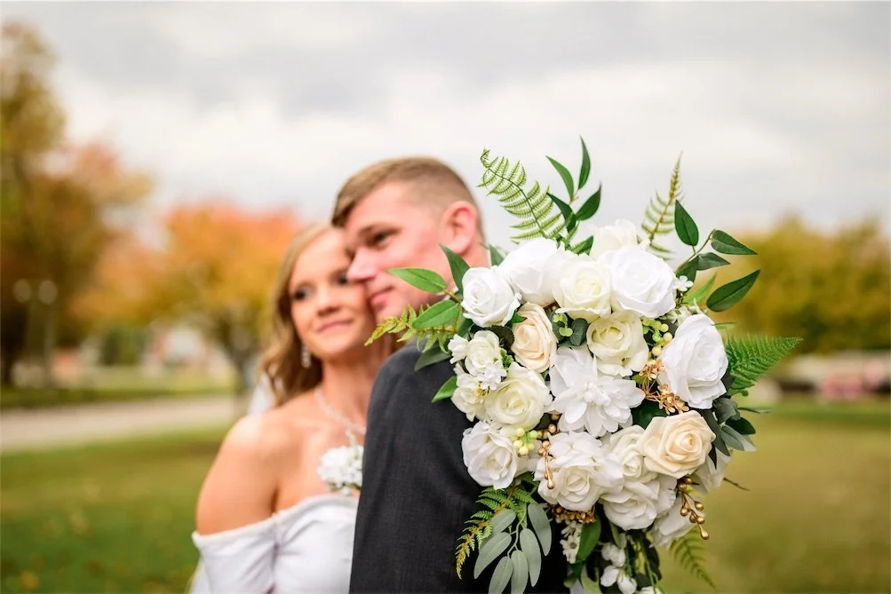 Close up of bride and groom cheek to cheek behind a white rose bouquet with autumn trees blurred