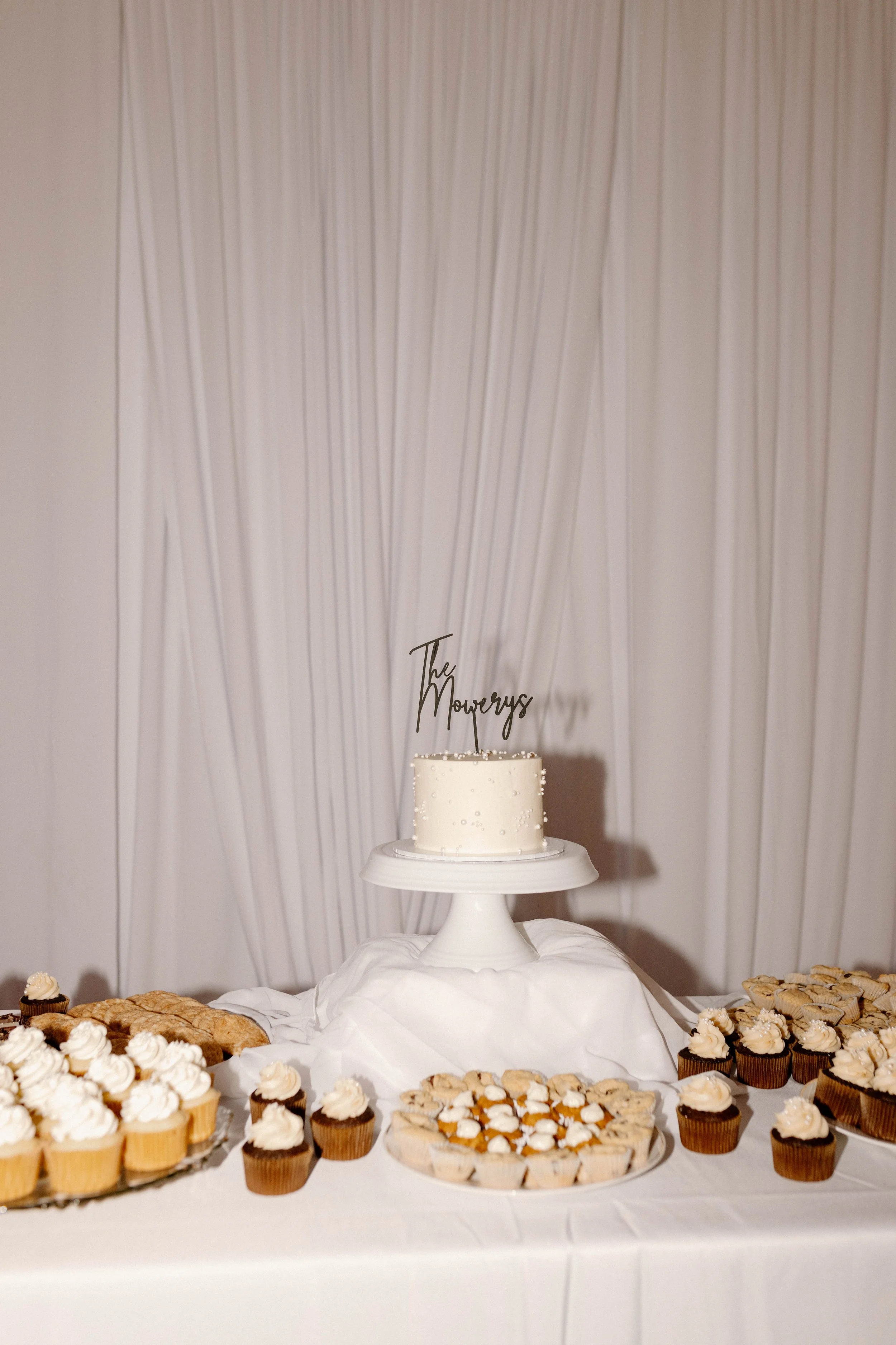 Wedding dessert table with a white cake on a cake stand, decorated with topper reading 'The Mowerys', surrounded by cupcakes and cookies.