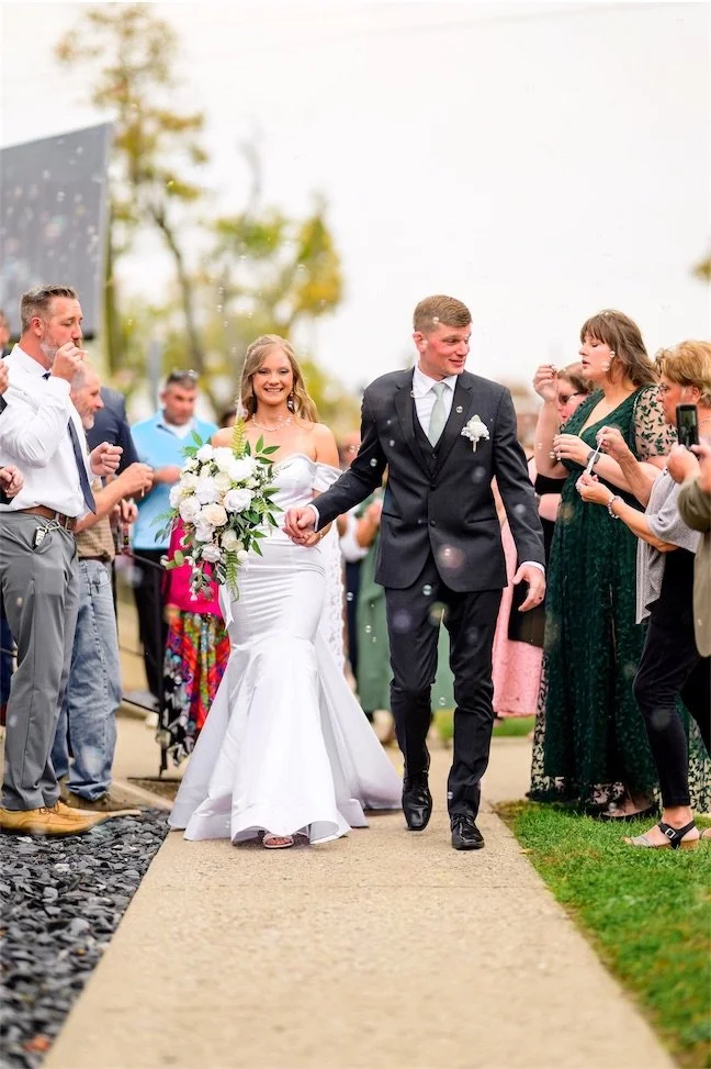 Bride and groom walk down a sidewalk lined with cheering guests after their ceremony