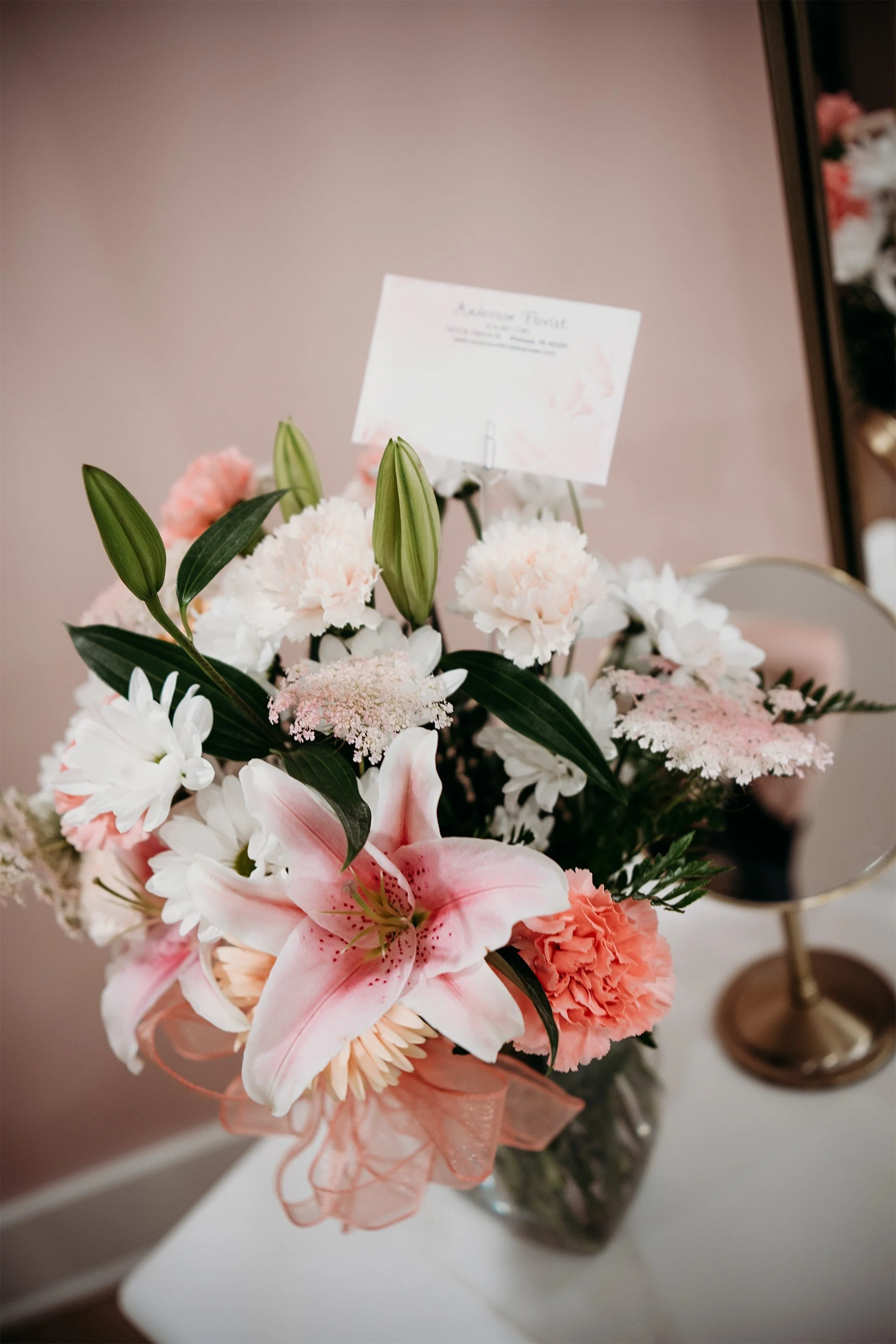 Pink and white lily and carnation bouquet in a glass vase beside a gold framed mirror in the bridal suite
