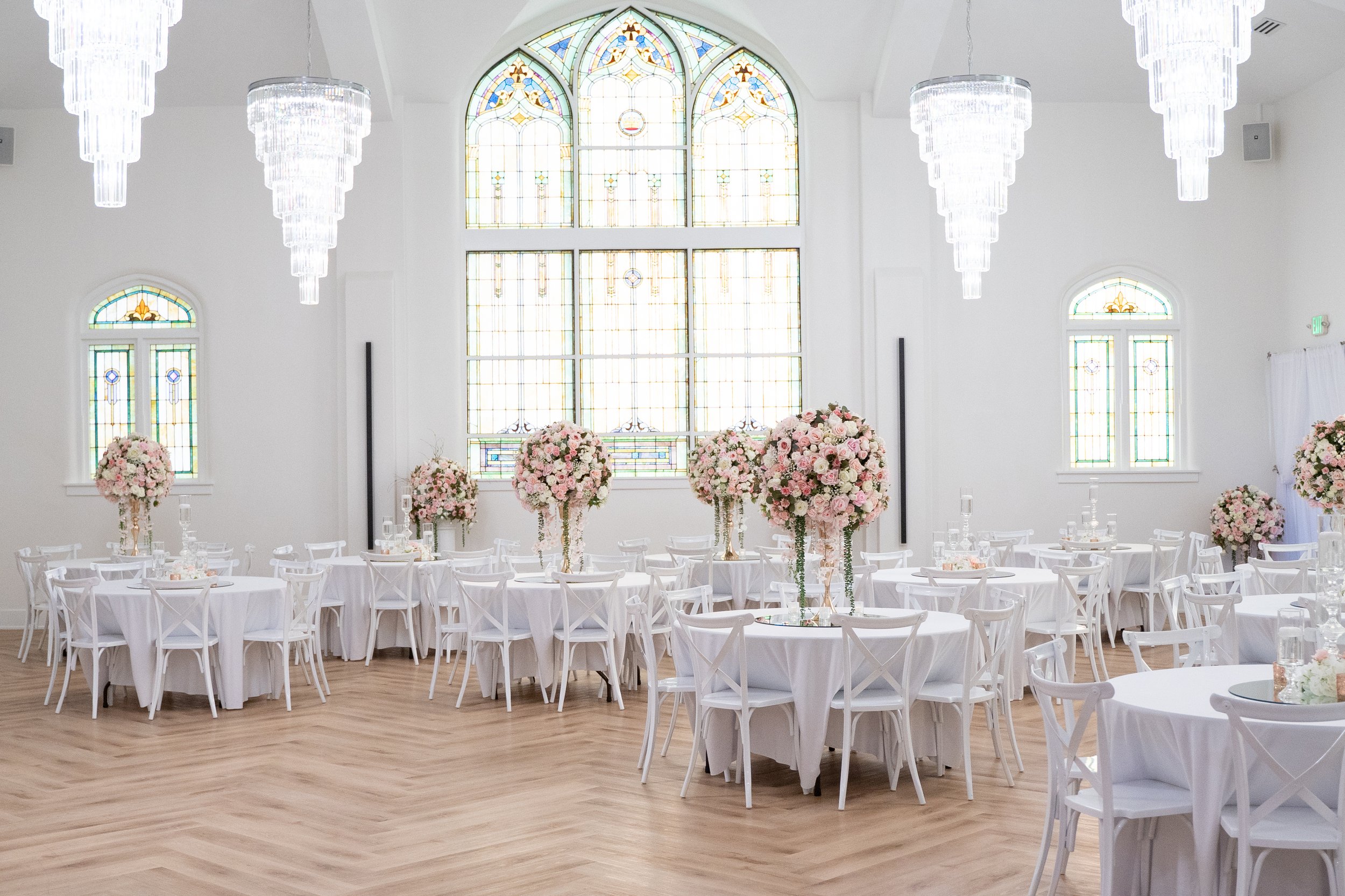 Elegant white wedding reception venue with crystal chandeliers, stained-glass windows, round tables, white cross-back chairs, and tall blush-pink rose floral centerpieces