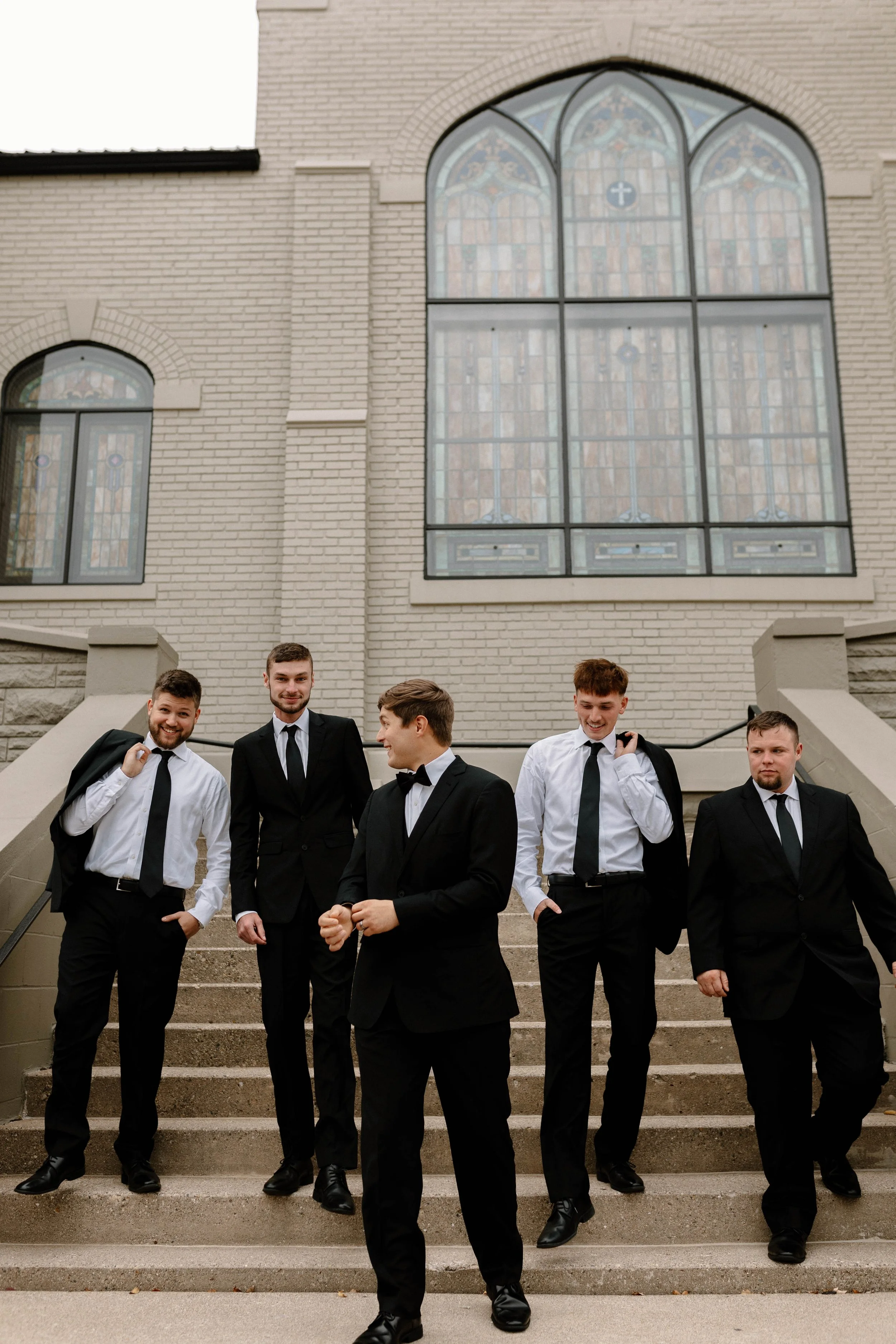 A group of five young men dressed in formally black suits and white shirts, with some carrying black jackets over their shoulders, standing on the steps of a church outside a large stained glass window.