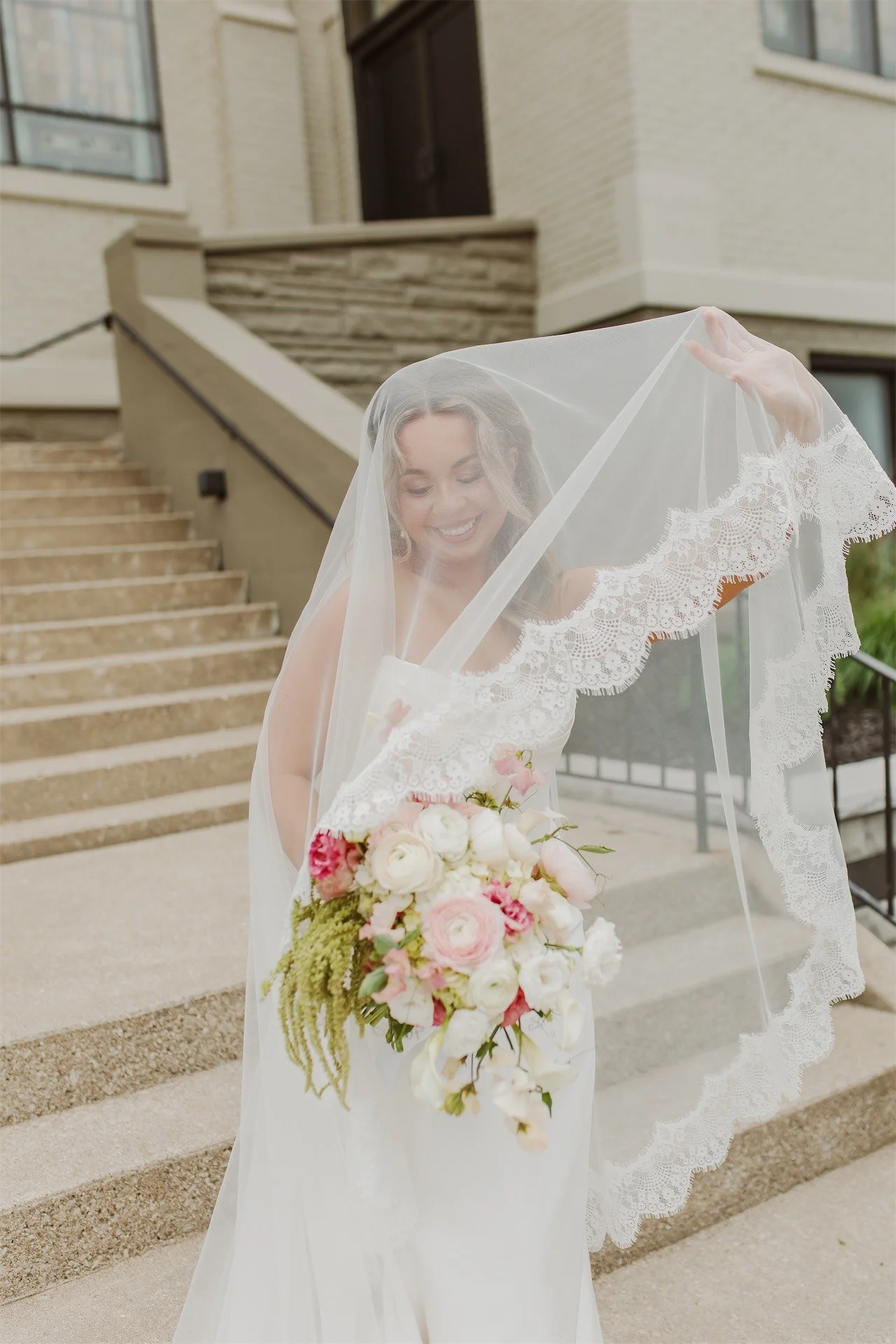 Bride laughing while lifting lace-trimmed cathedral veil on church staircase