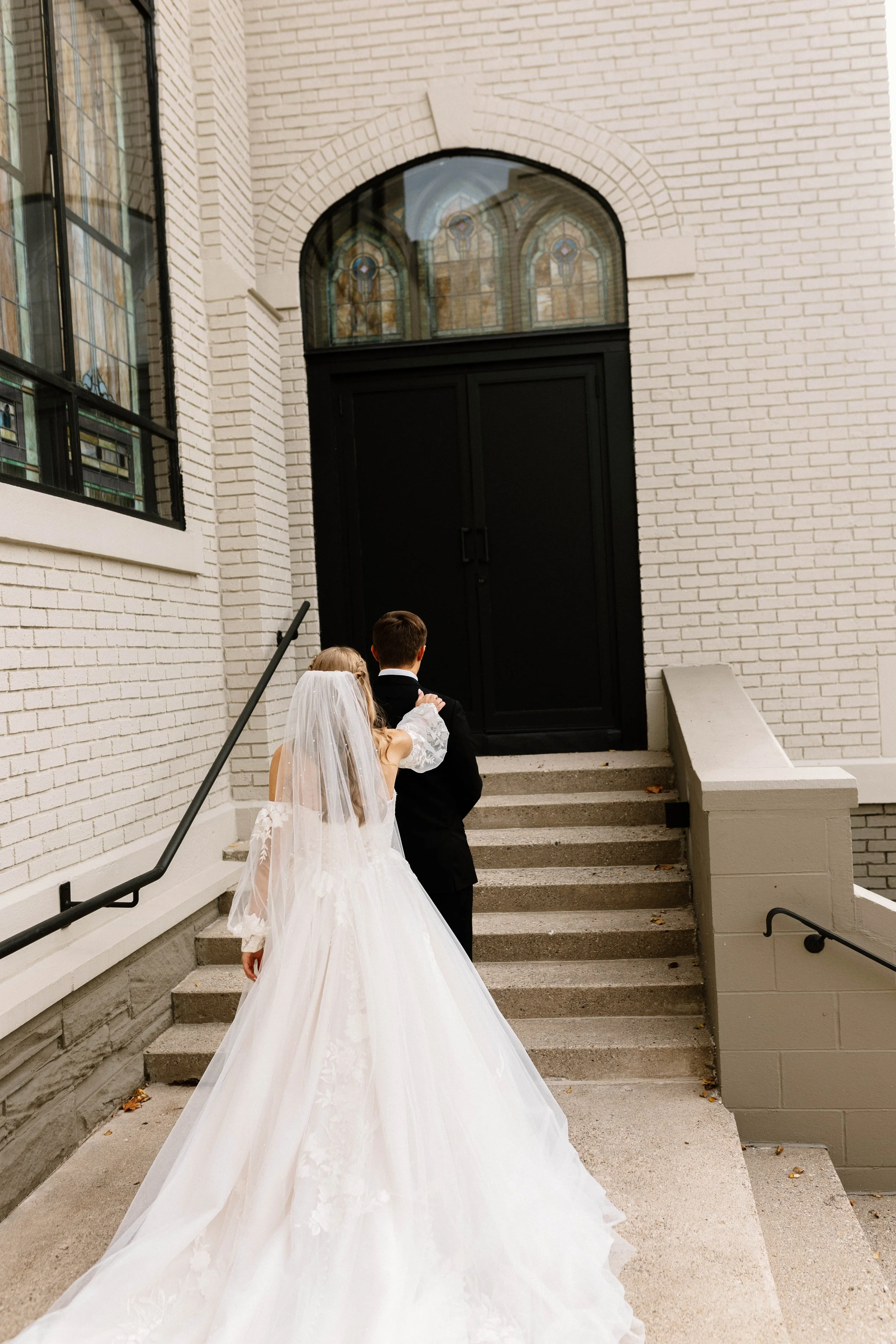 A bride in a white wedding gown and veil, and a groom in a black suit, ascending steps outside a building with white brick walls and stained glass windows, heading toward a black door.