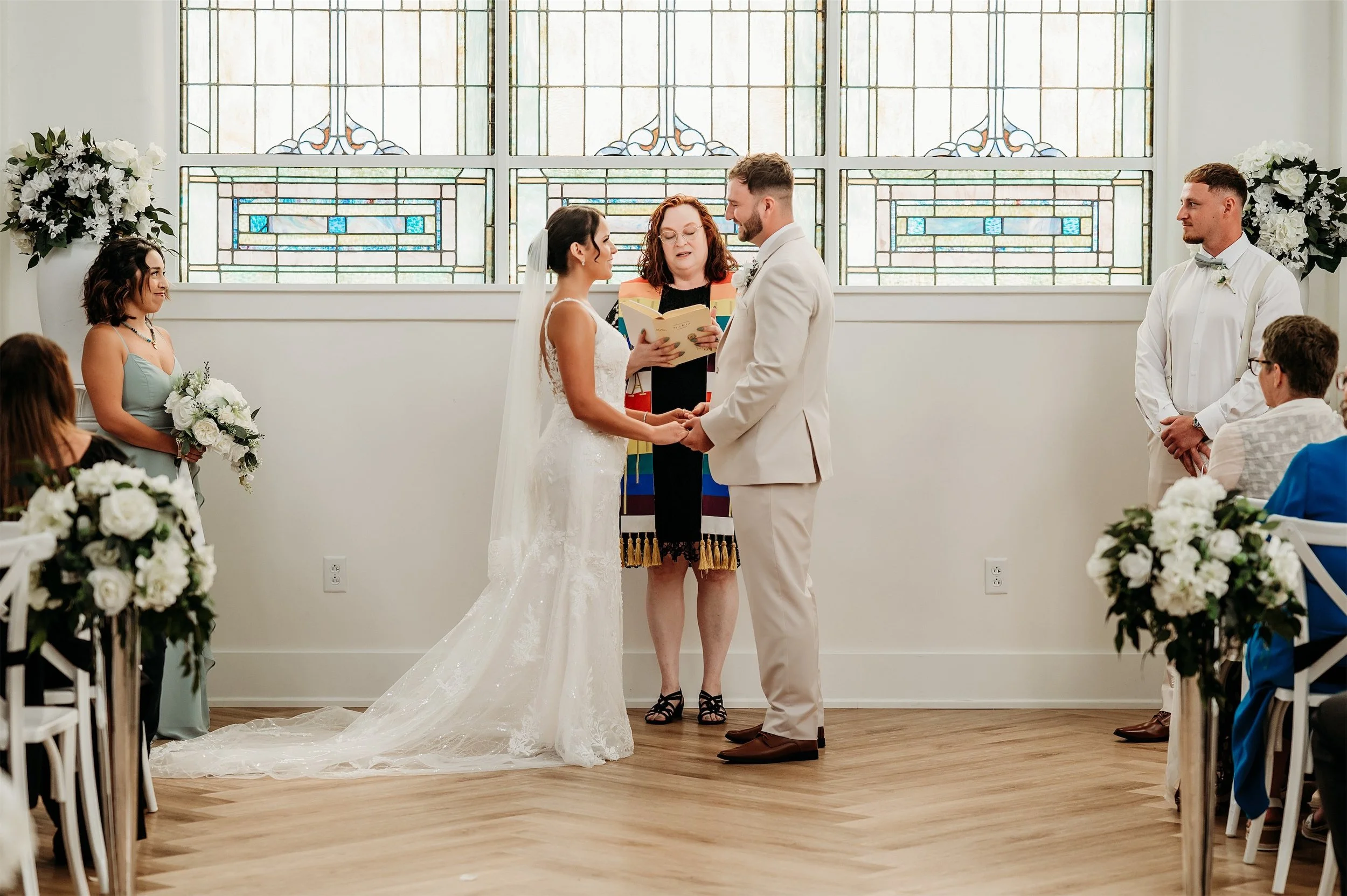 Bride and groom holding hands during their ceremony in front of stained glass windows with an officiant