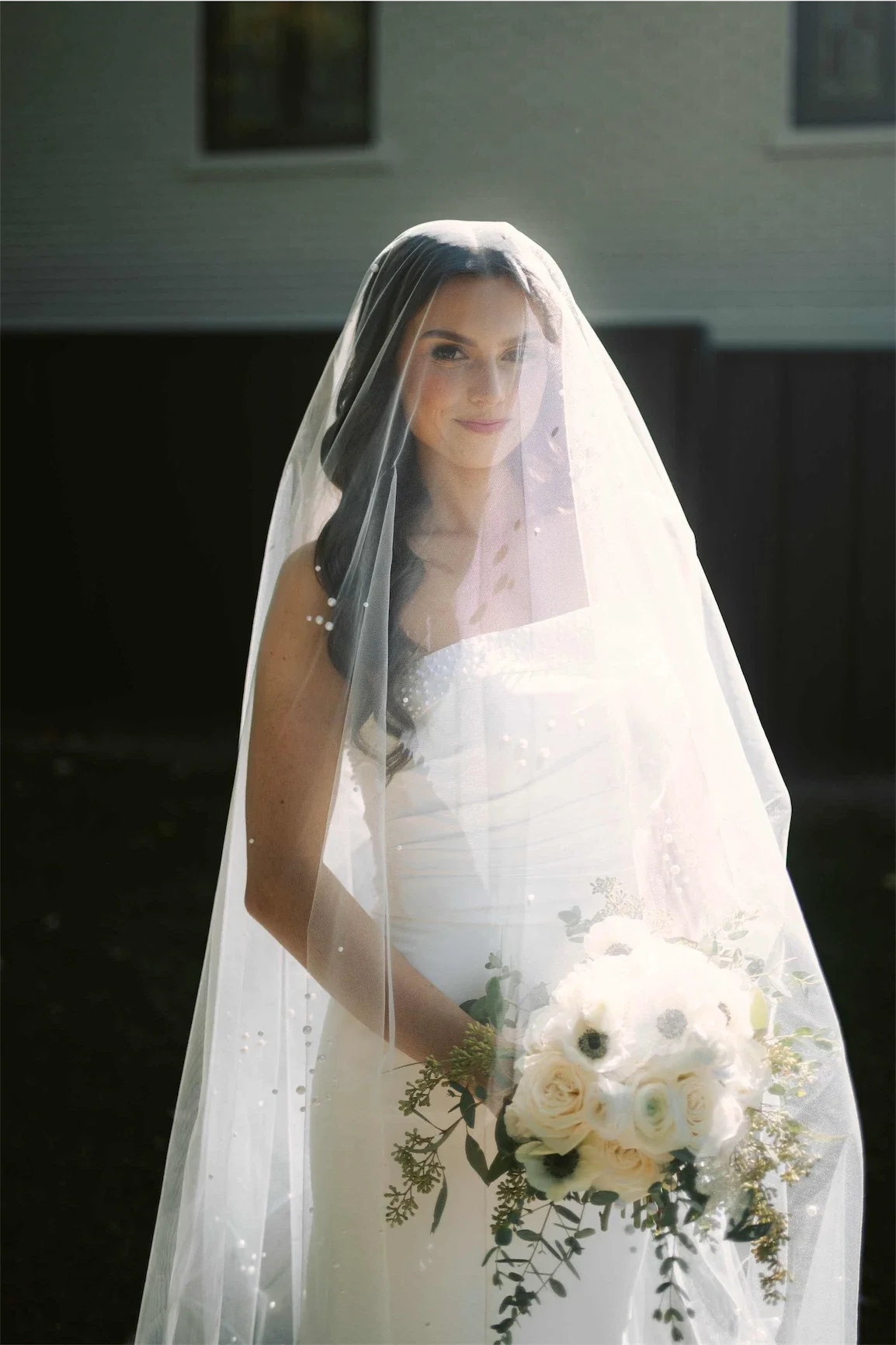 Bride smiling softly beneath her pearl veil while holding a white rose and anemone bouquet