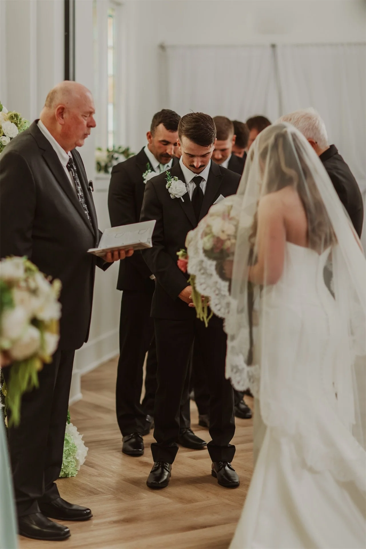 Groom teary eyed listening to officiant during vows beneath stained glass window