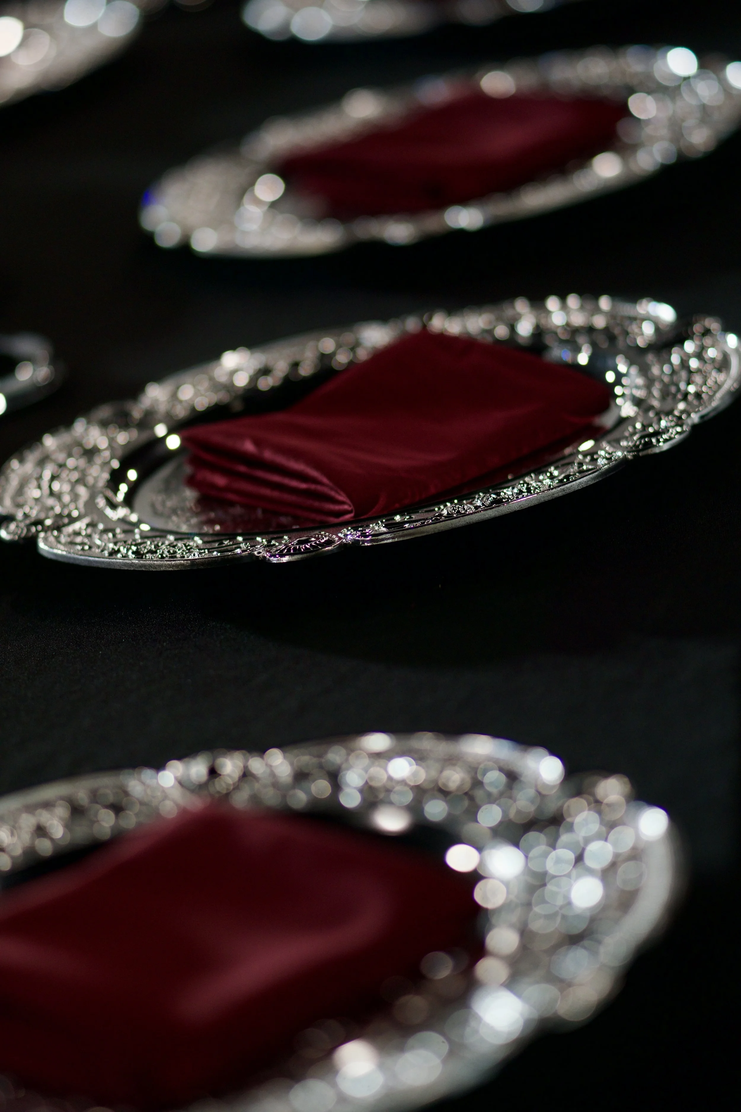 row of ornate silver chargers with burgundy napkins set on black table linen