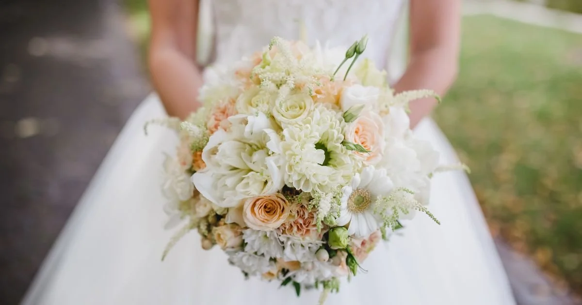 Close-up of summer wedding bouquet with peach roses, white dahlias, and greenery held by bride