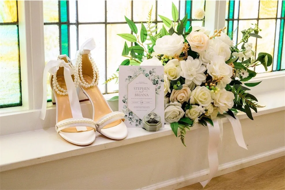 Pearl strap bridal heels, wedding invitation and white rose bouquet on a stained glass windowsill