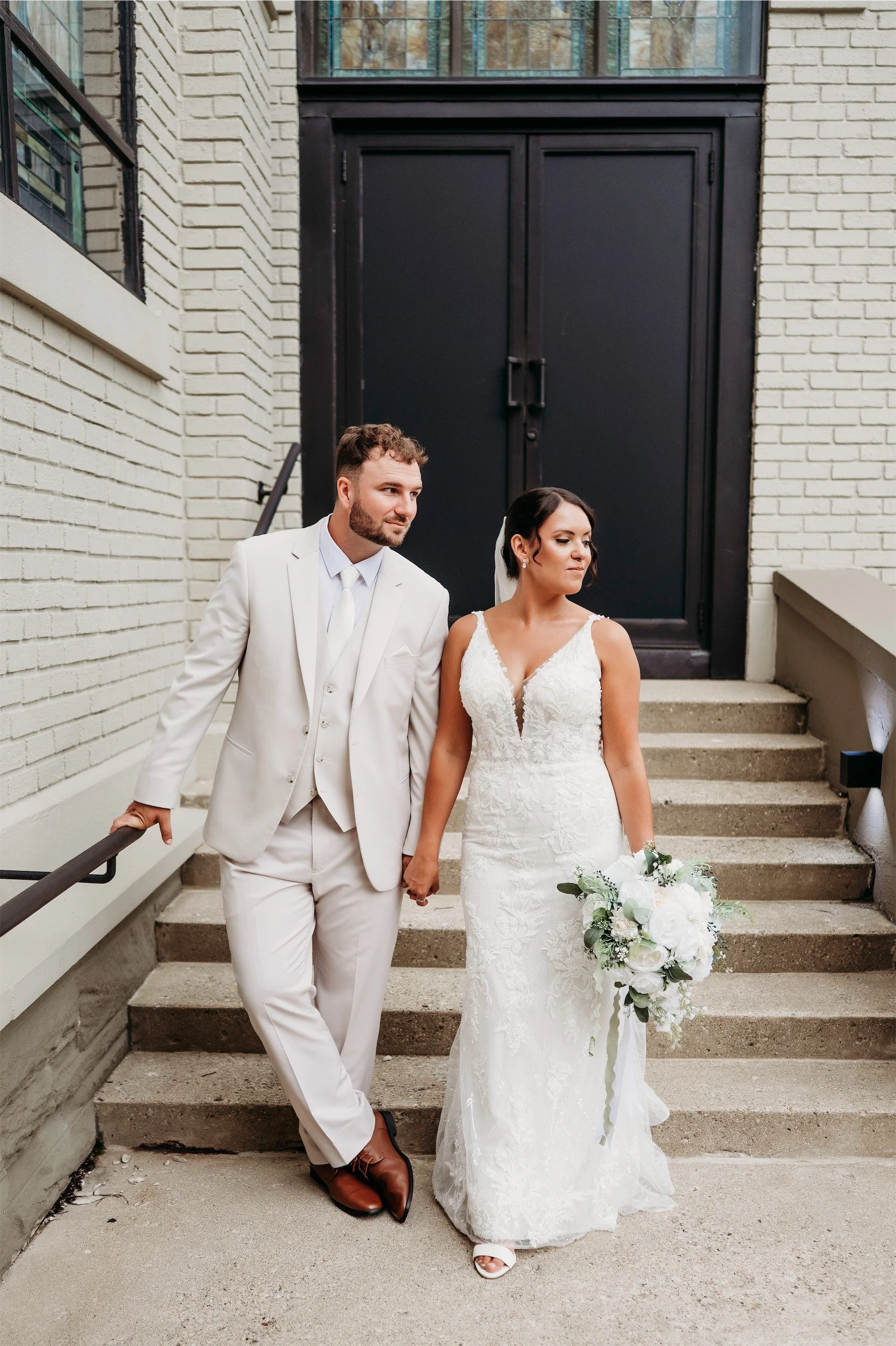Couple holding hands in front of black double church doors on brick steps with the bride carrying a white bouquet