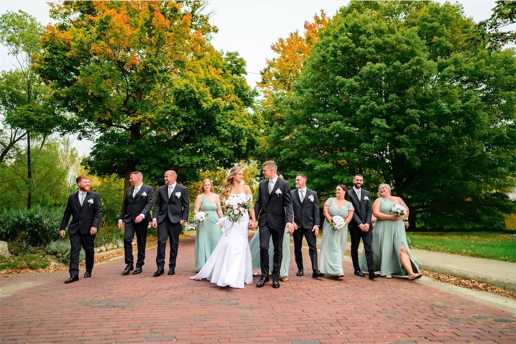 Bridesmaids, groomsmen and the newlyweds walk together down a brick path beneath colorful fall trees
