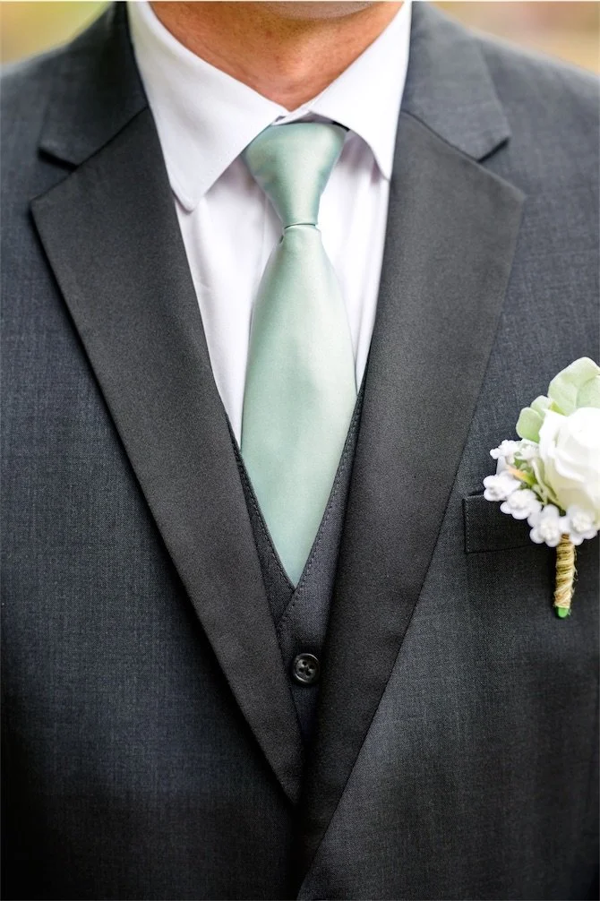 Close up of a groom's sage green tie, charcoal vest and white rose boutonniere
