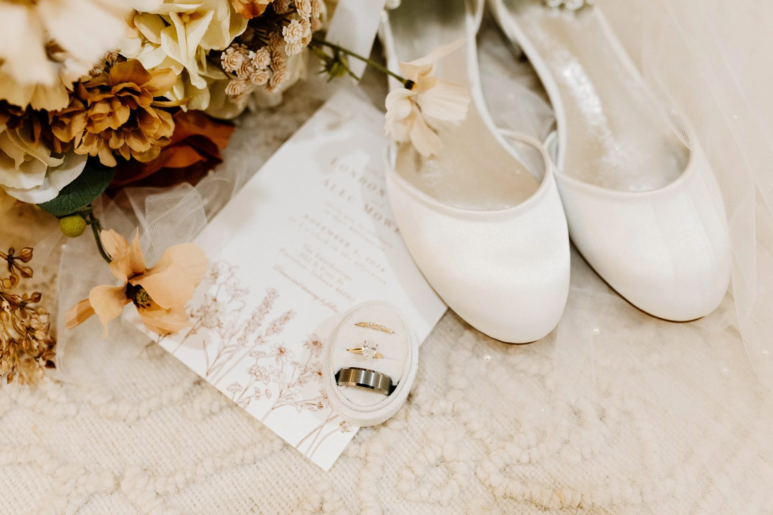 A pair of white wedding shoes, a bouquet of dried flowers, a wedding invitation, and a set of wedding rings in a small white box on a lace fabric surface.