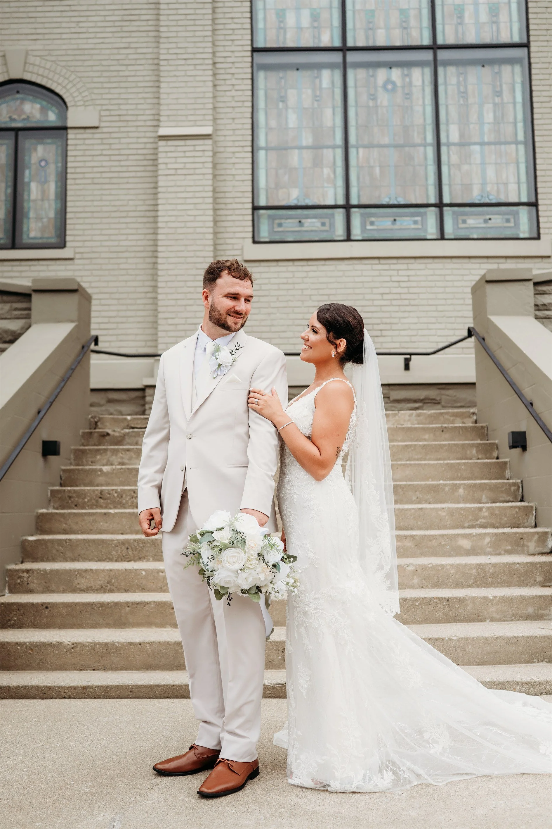 Bride and groom looking at each other on the brick church steps with a white floral bouquet