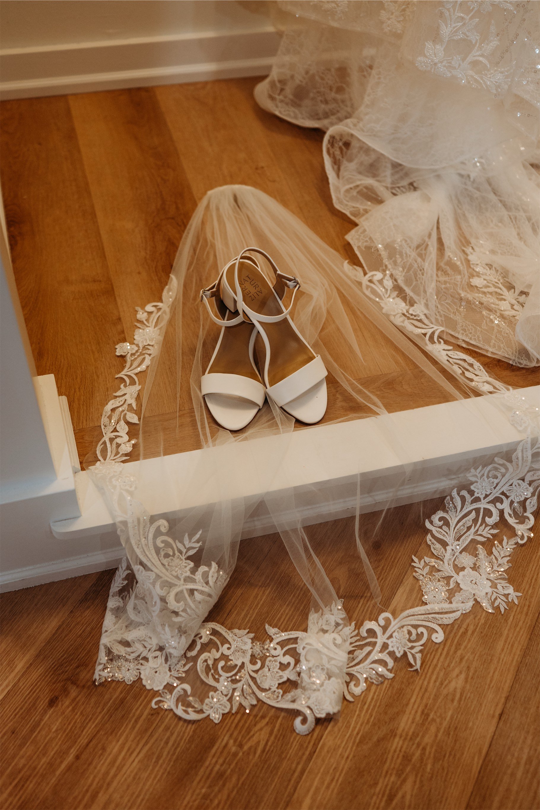 White strappy bridal sandals resting on a lace trimmed veil on a wood floor