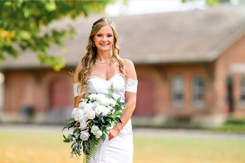 Smiling bride holds a white and green bouquet in front of a blurred red brick building
