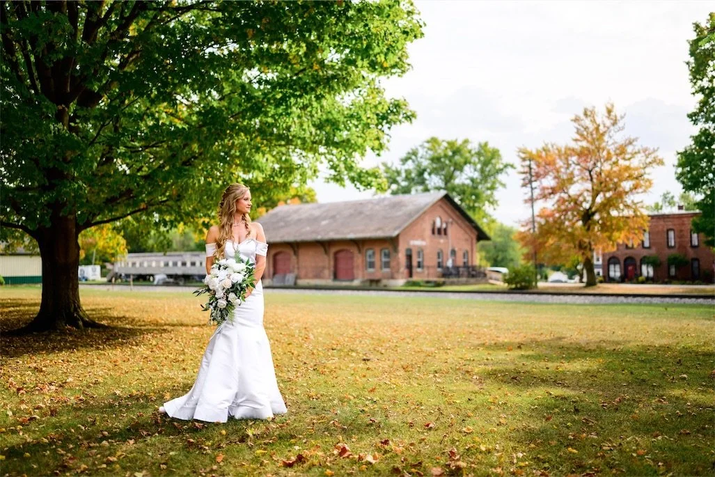 Bride in a fitted white gown holding a bouquet under a large tree on an autumn lawn
