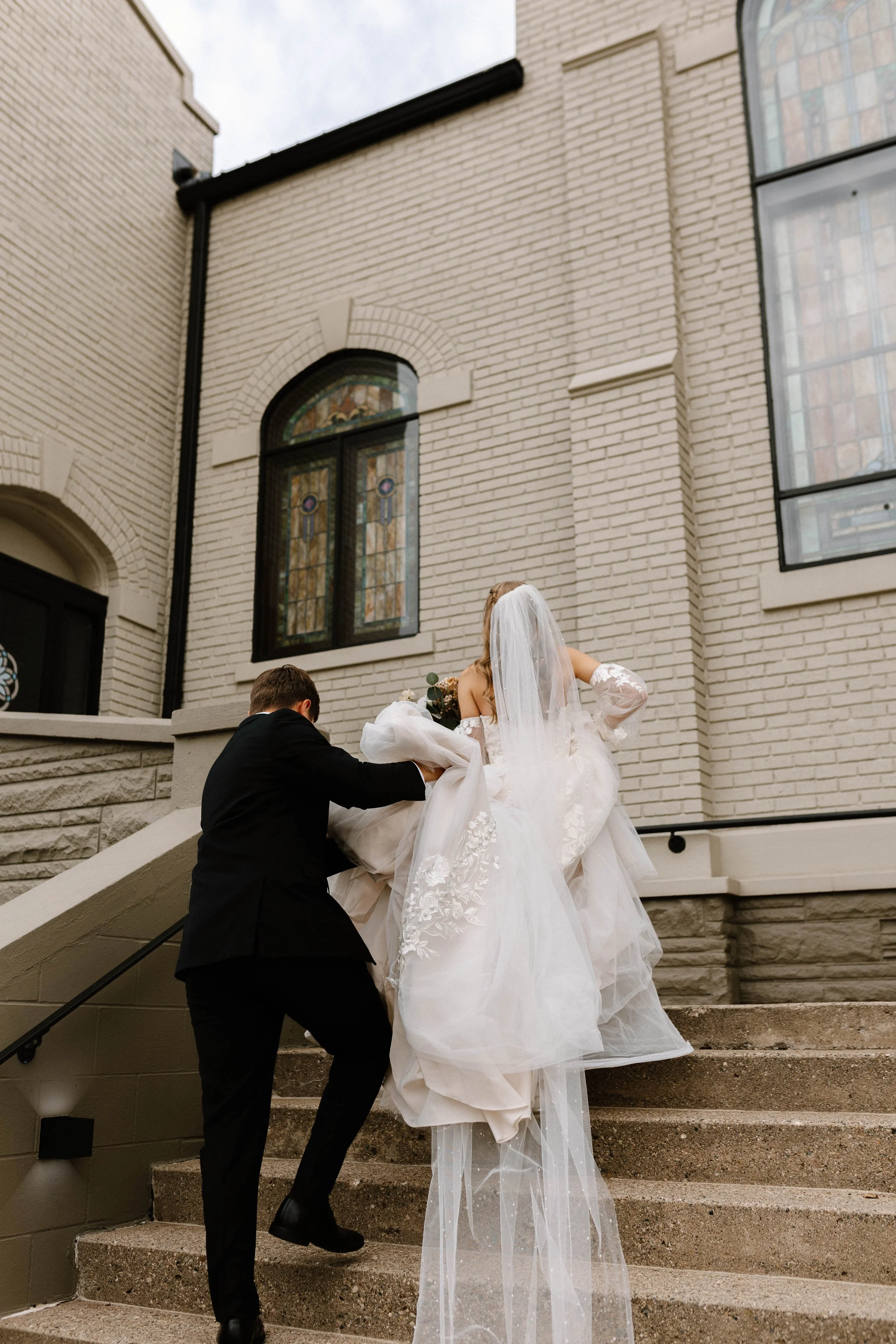 A groom helping a bride in a white wedding gown up the steps of a church.