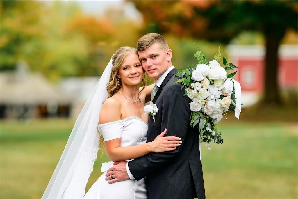Bride and groom embrace and smile at the camera holding a white bouquet on an autumn lawn