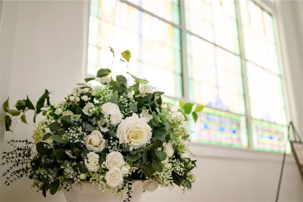 Large white rose and greenery arrangement in front of a tall arched stained glass window