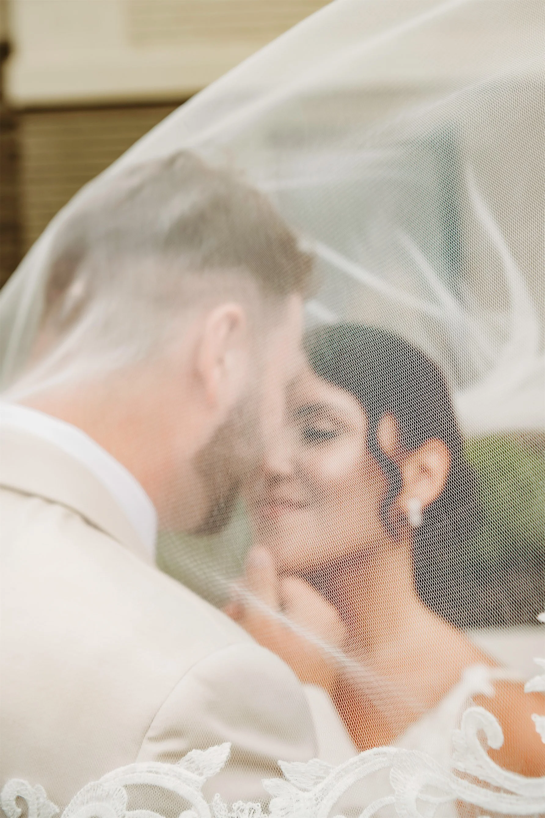 Groom leaning in to kiss the smiling bride behind a sheer veil caught in the breeze