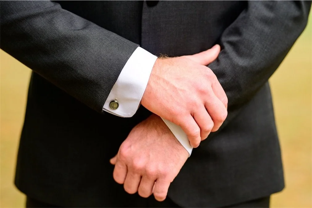 Groom's crossed hands showing a gold cufflink and white shirt cuff against a charcoal suit