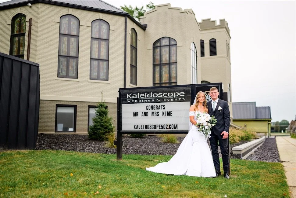 Newlyweds pose beside the Kaleidoscope Weddings and Events congratulations sign outside the Indiana venue