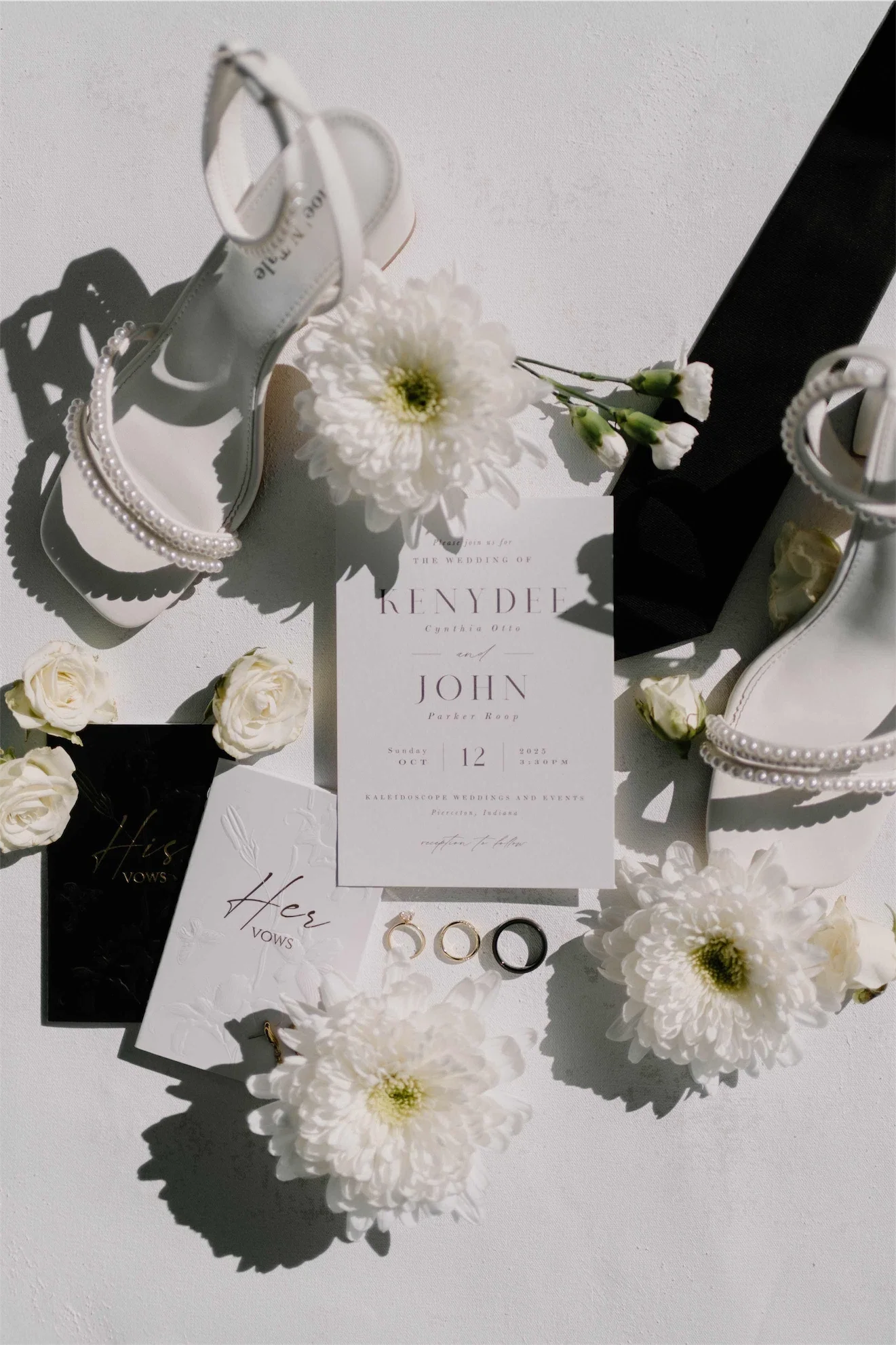 Minimalist wedding invitation surrounded by pearl strap heels, his and her vow books, rings, and white chrysanthemums