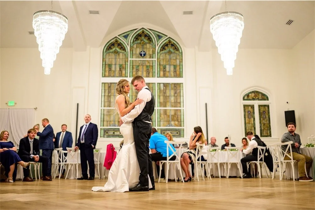Bride and groom share a slow dance on the wood floor while guests sit at tables behind them