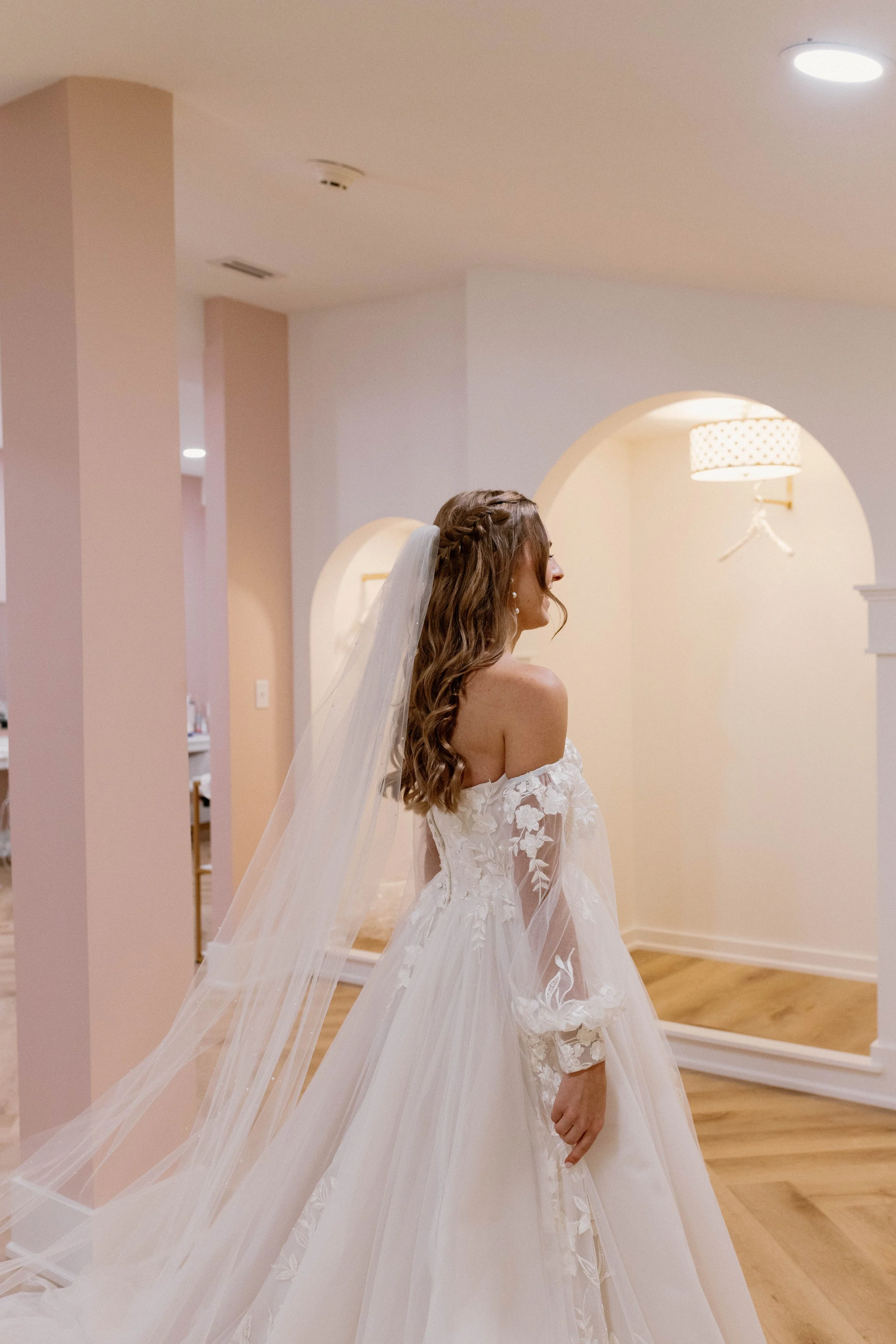 A bride in a white off-the-shoulder wedding dress with lace details and a long veil, standing in a softly lit indoor space with light-colored walls and wooden flooring.