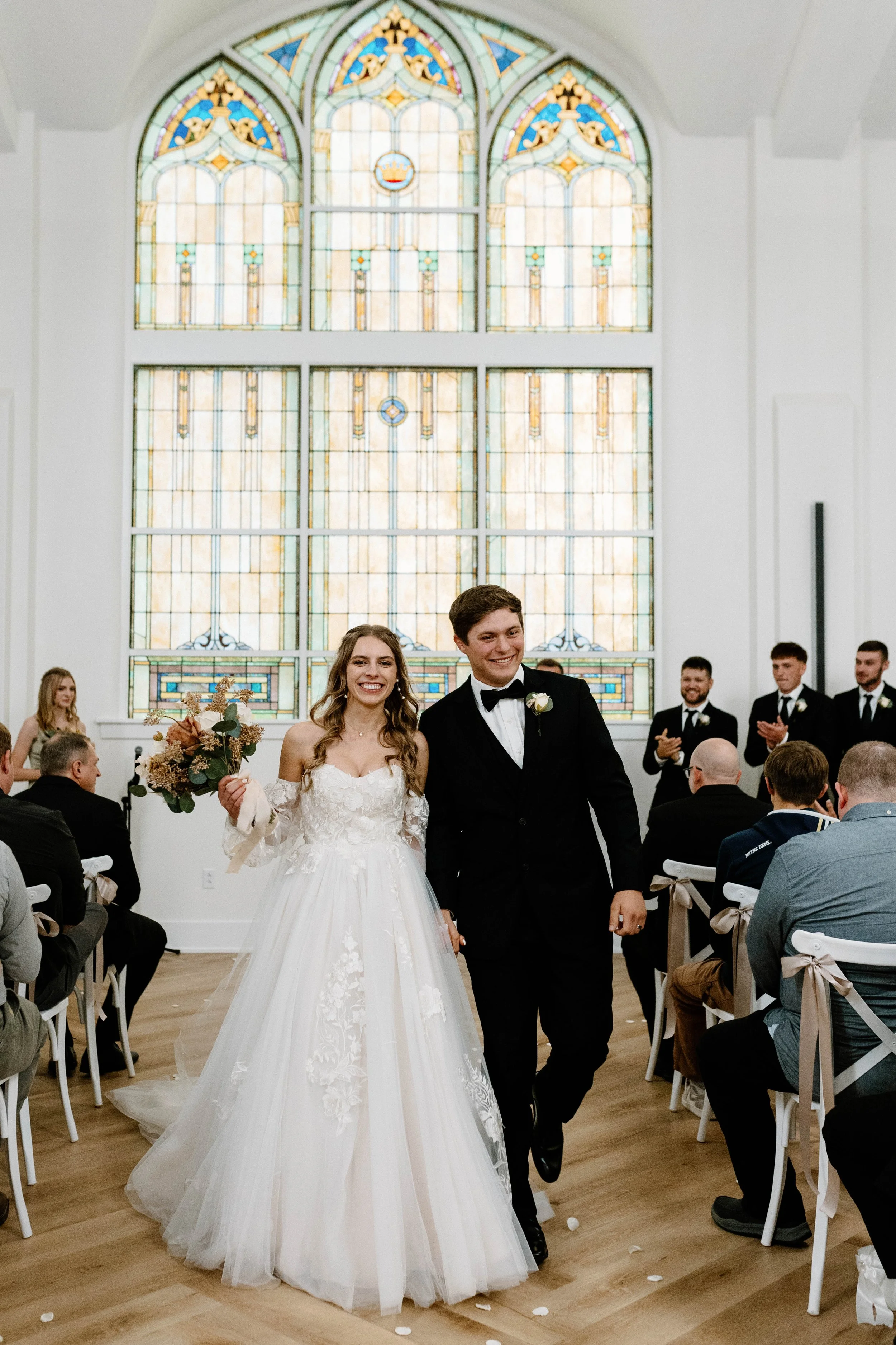 A newlywed couple walking down the aisle in a church after their wedding ceremony, surrounded by seated guests, with a stained glass window in the background.