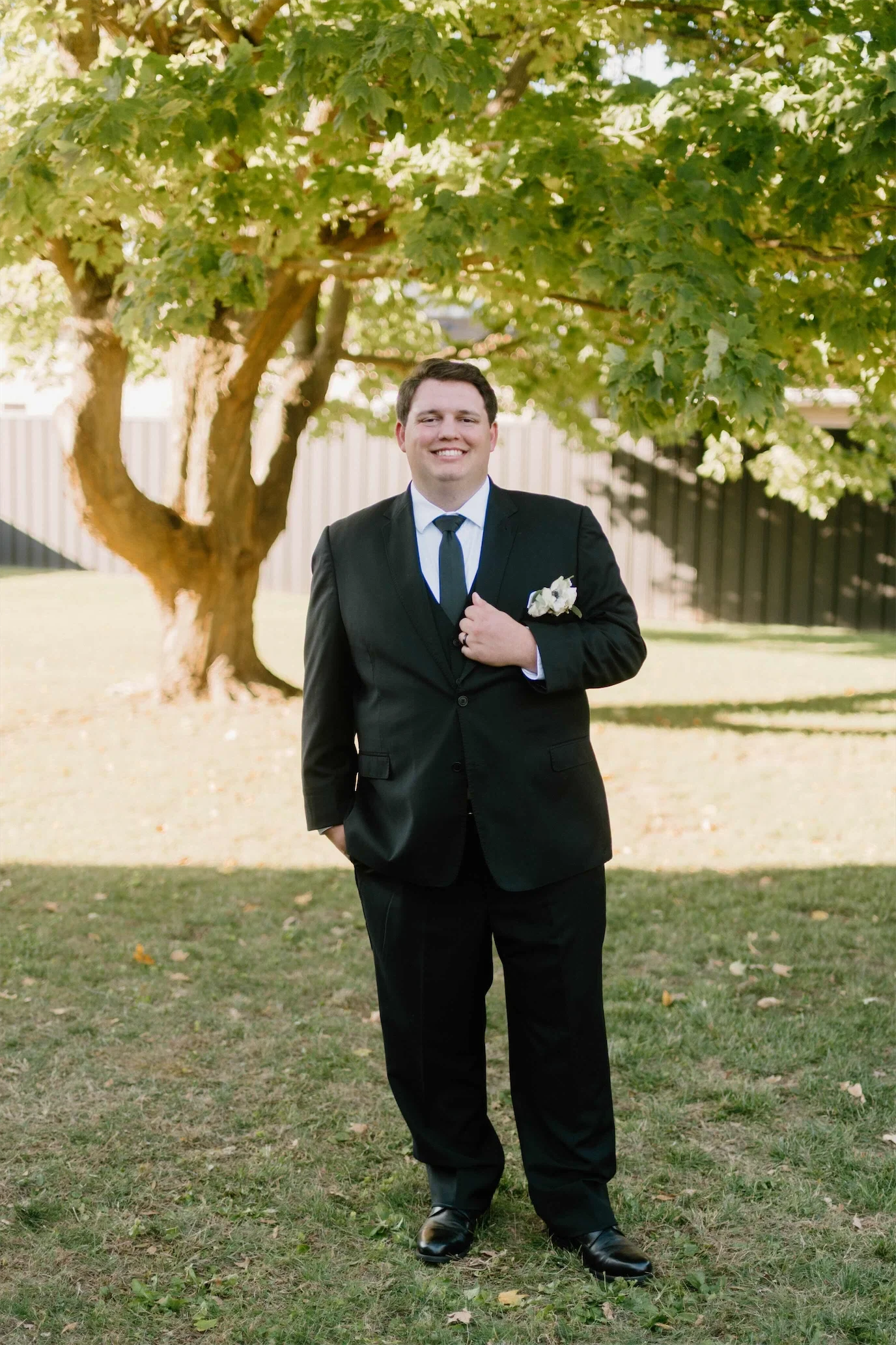 Smiling groom in a black suit and white boutonniere standing under a shade tree