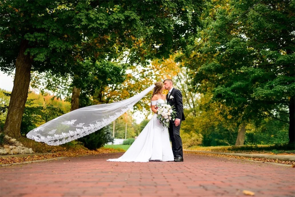 Bride's lace veil flows in the wind as the couple kiss on a brick path under autumn trees