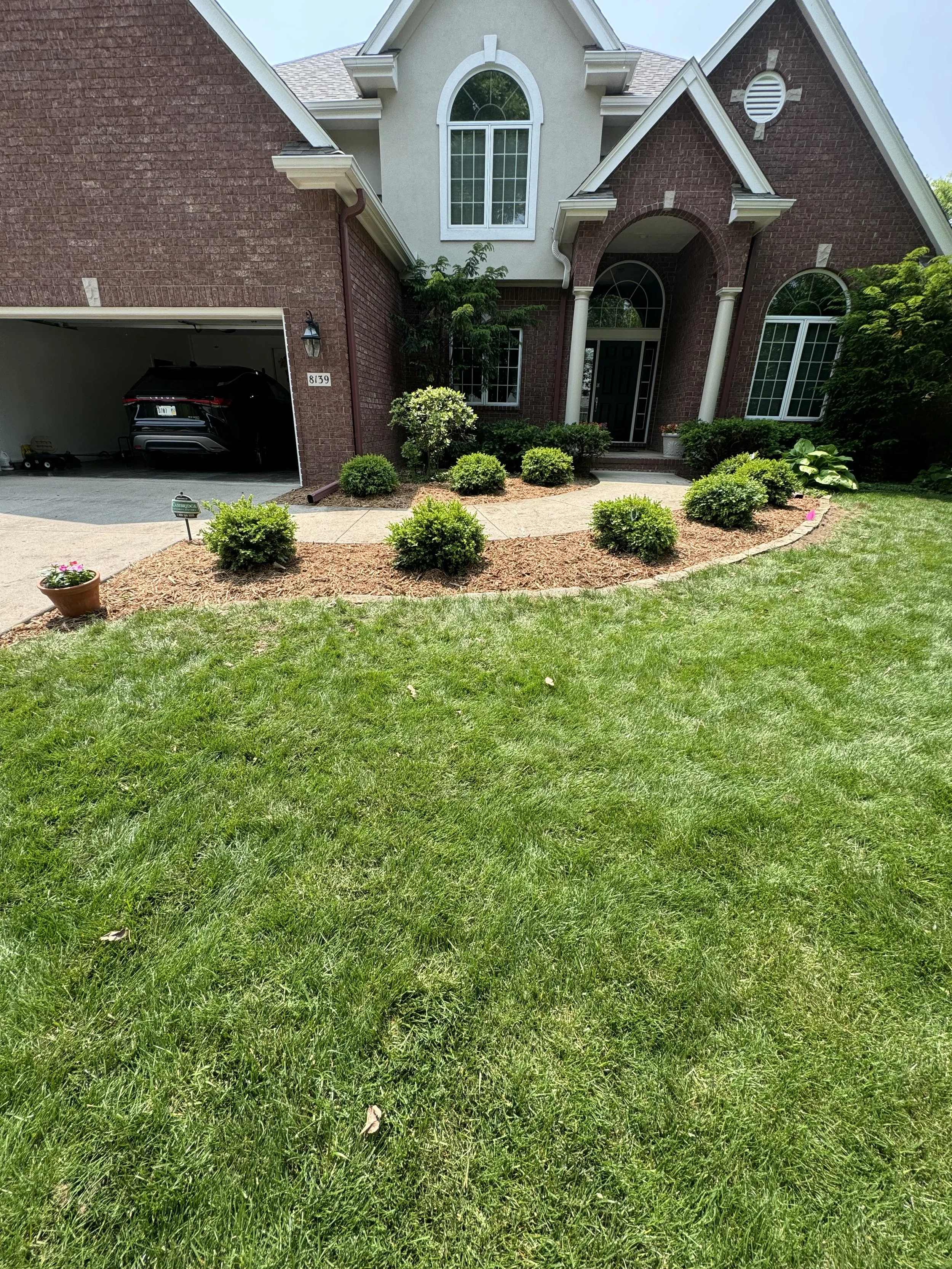 Front yard of a brick house with a well-maintained lawn, a garden bed with small bushes, and a concrete pathway leading to the front door. The house has large windows, columns at the entrance, and a garage to the left with a black vehicle inside.