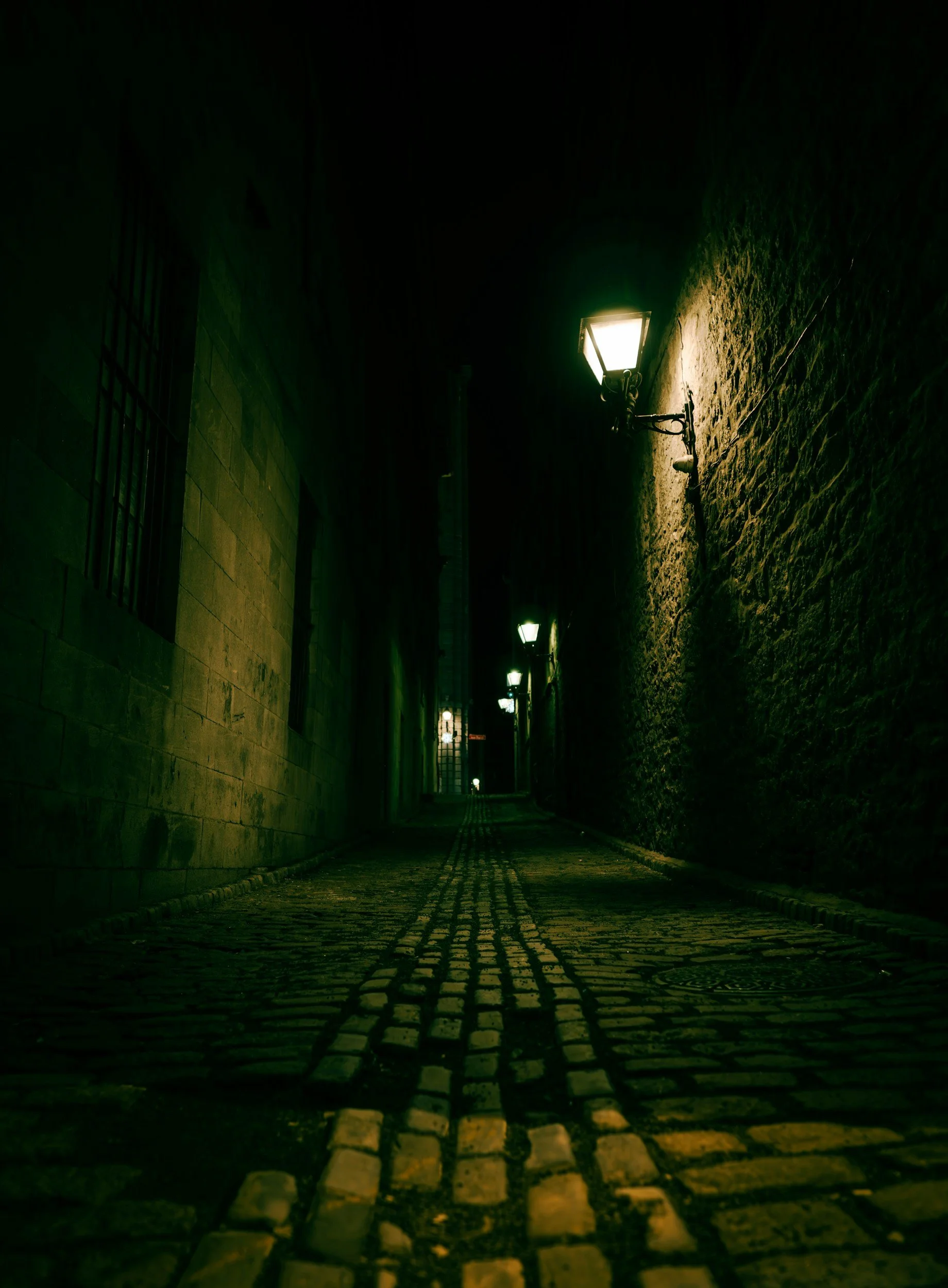 Nighttime scene of a narrow cobblestone alleyway illuminated by old-fashioned street lamps attached to brick walls.