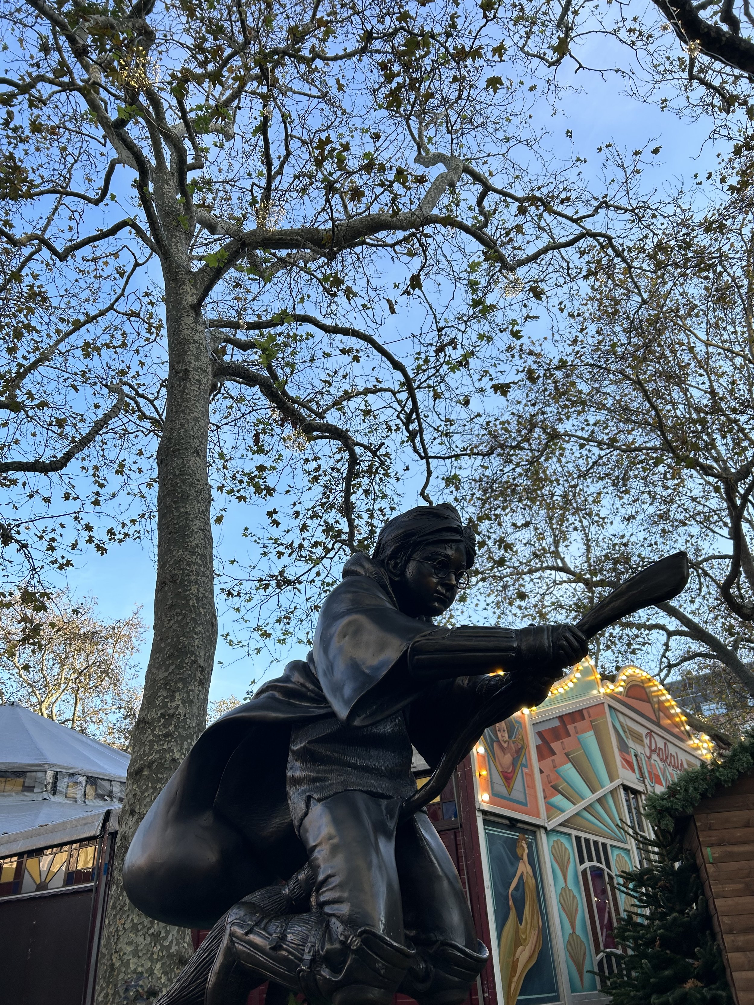 A bronze statue of a Harry Potter on Leicester Square, wearing glasses and a coat, set outdoors among leafless trees with a blue sky and carnival booths in the background.