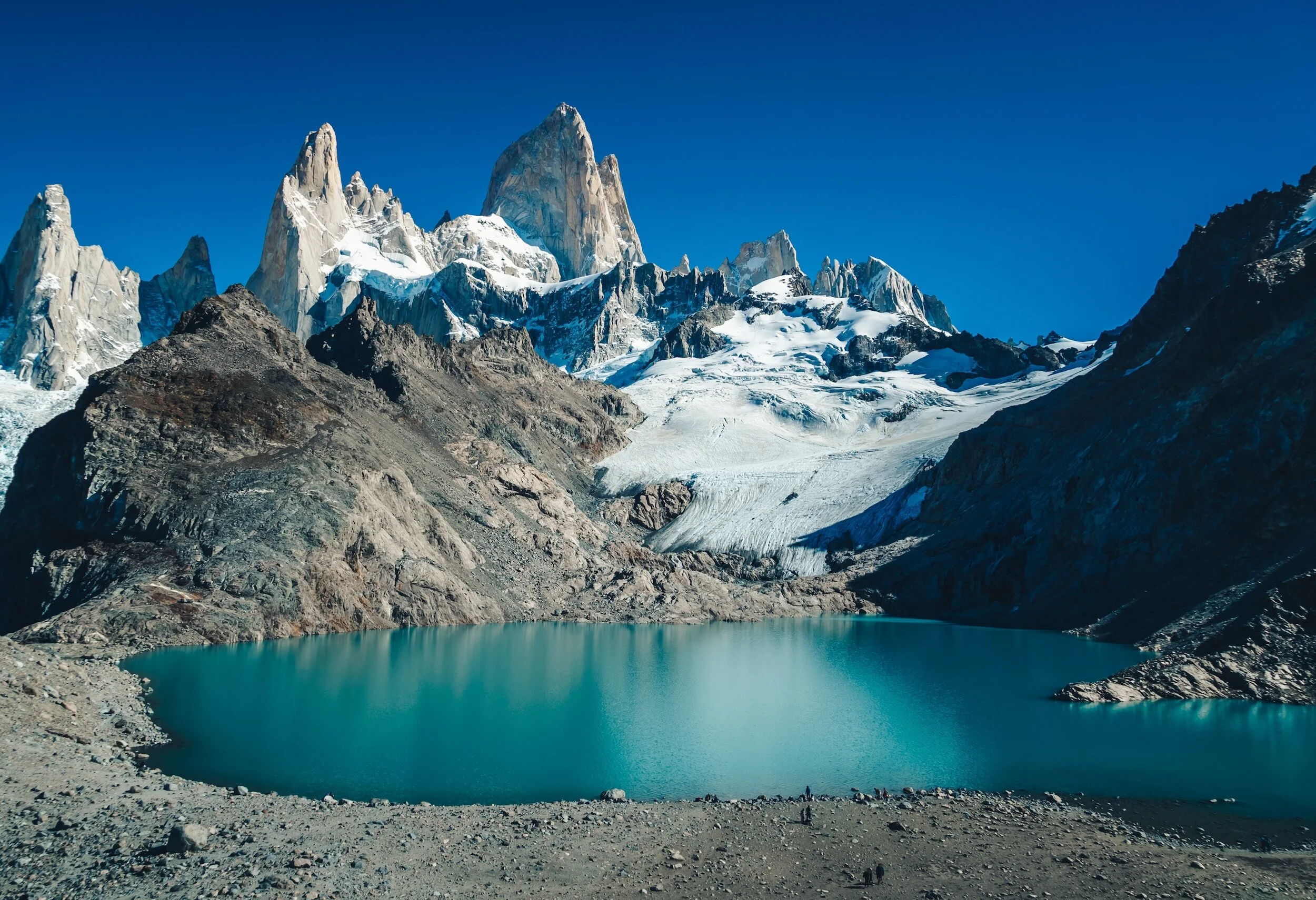Snow-capped mountains with a glacier, a teal lake in the foreground, and a clear blue sky.