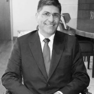 A black-and-white photo of a man in a suit and tie, smiling and sitting at a table indoors.