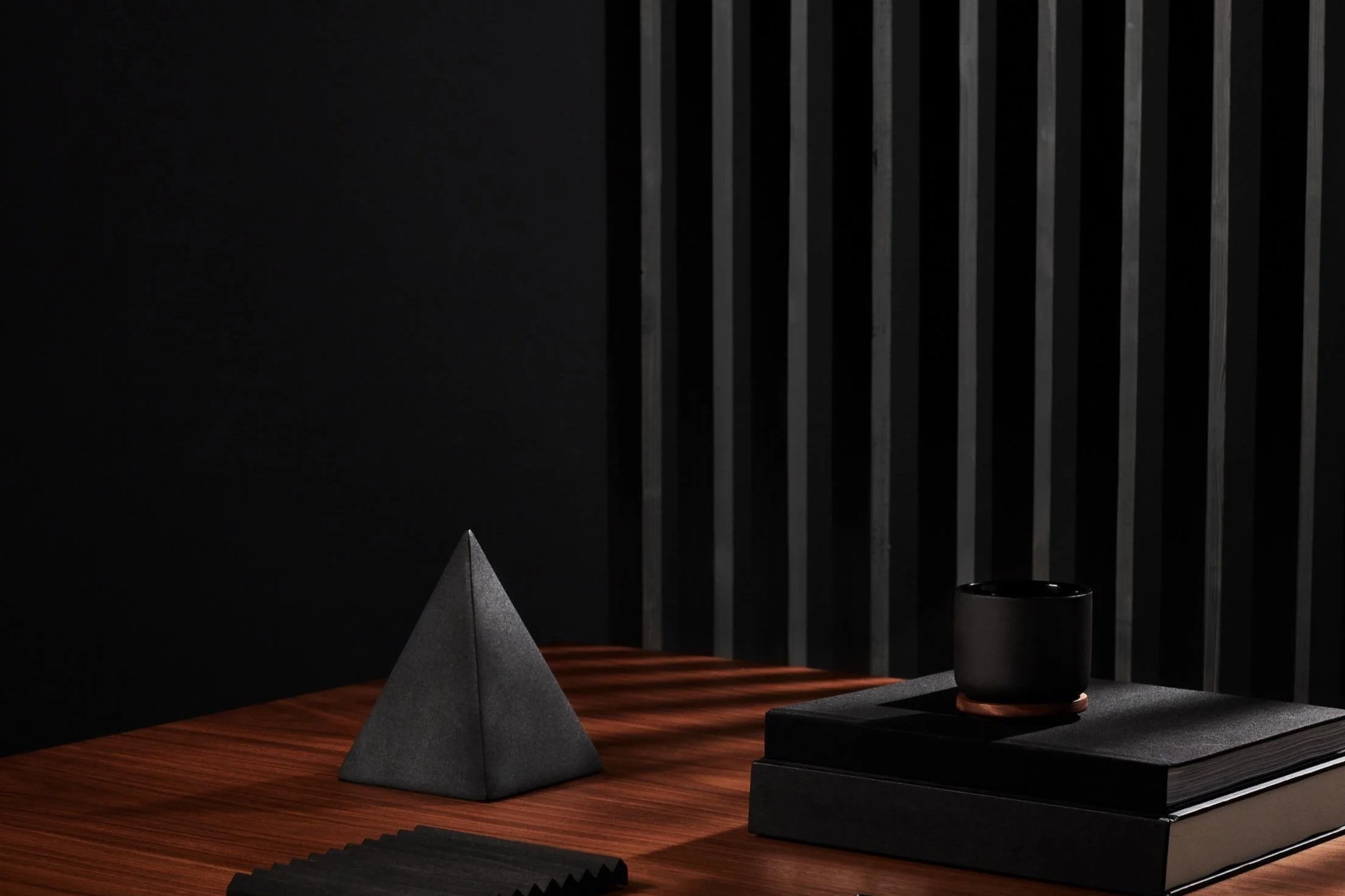 Dark wooden desk with a black pyramid sculpture, a black cup, and a stack of black notebooks against a black wall with vertical black slats.